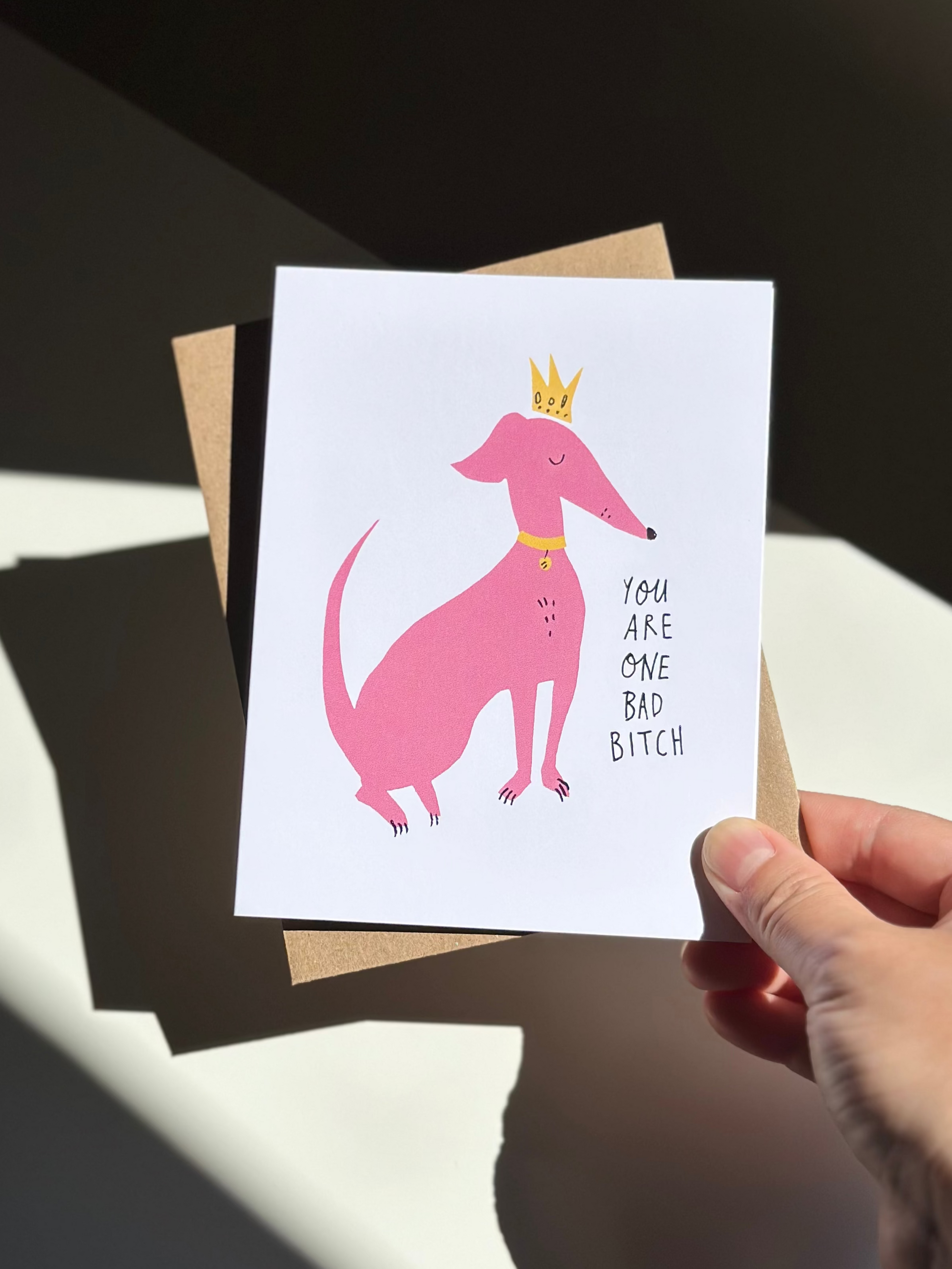 Funny greeting card with pink cartoon dog wearing a yellow crown and collar, captioned ‘You are one bad bitch,’ on white background with brown kraft envelope.