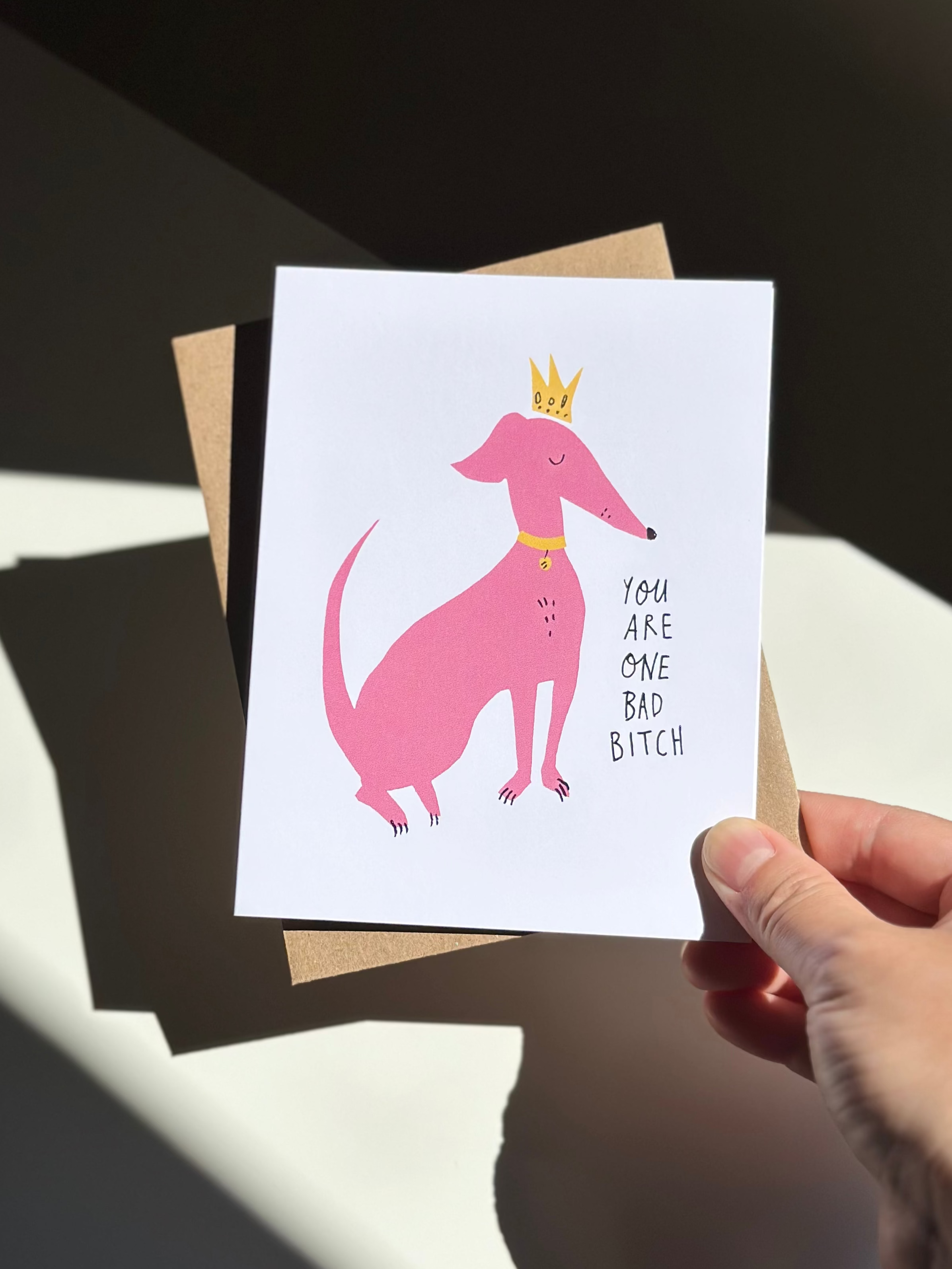 Funny greeting card with pink cartoon dog wearing a yellow crown and collar, captioned ‘You are one bad bitch,’ on white background with brown kraft envelope.