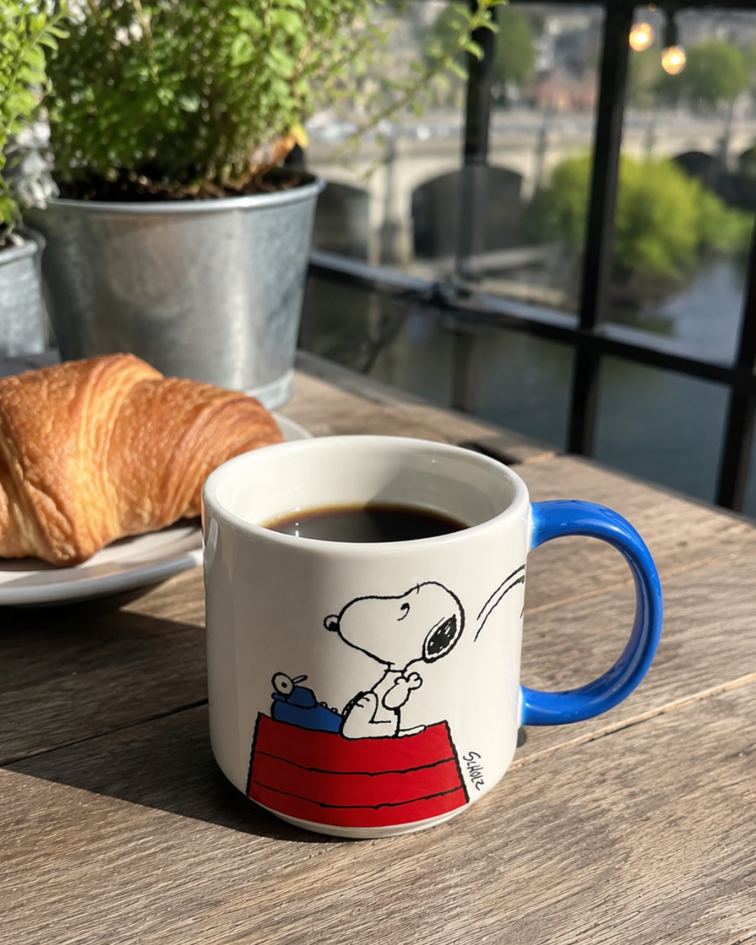 A blue-handled Snoopy mug featuring Snoopy typing on a typewriter atop his red doghouse sits on an outdoor café table by the water, beside a croissant and string lights.