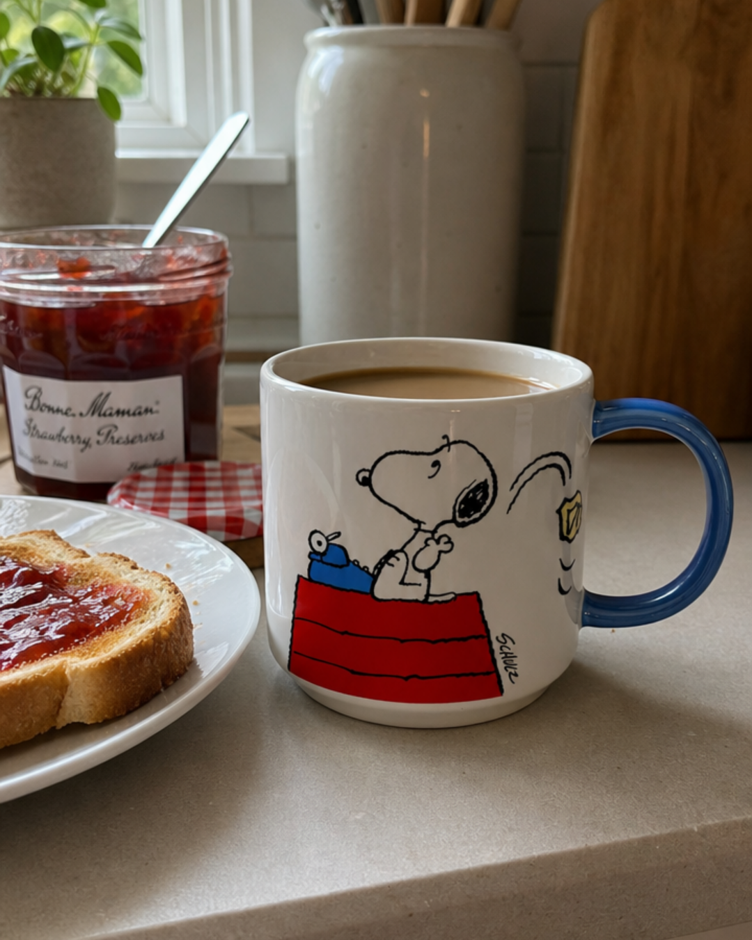 A blue-handled Snoopy mug featuring Snoopy typing on a typewriter atop his red doghouse sits on a kitchen counter beside toast with jam and a jar of strawberry preserves.