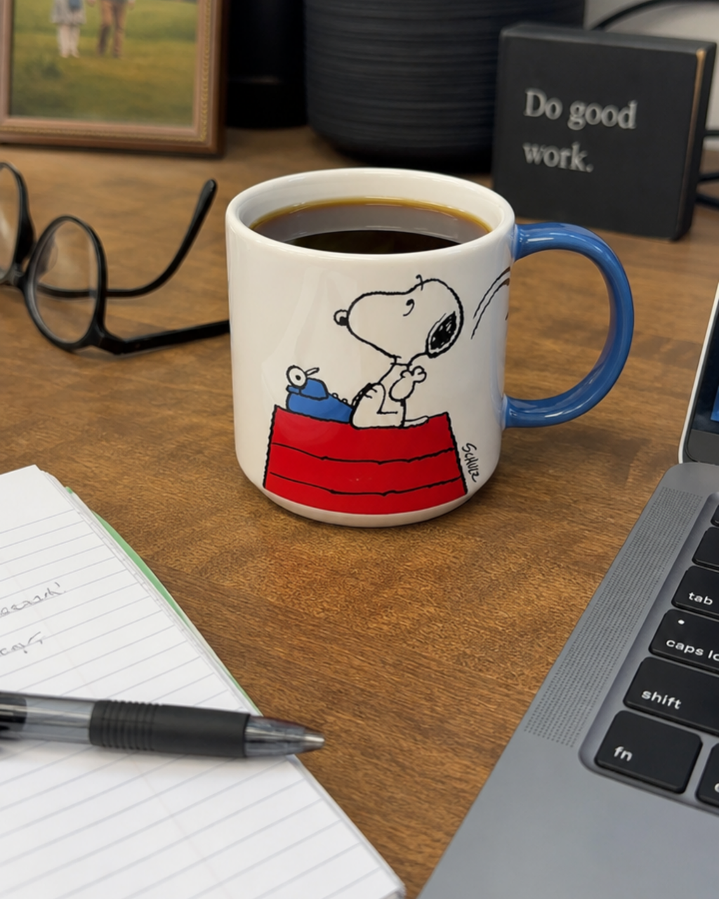 A blue-handled Snoopy mug featuring Snoopy typing on a typewriter atop his red doghouse with a crumpled page flying off, styled on a desk with glasses, a notepad, and a "Do good work" sign.