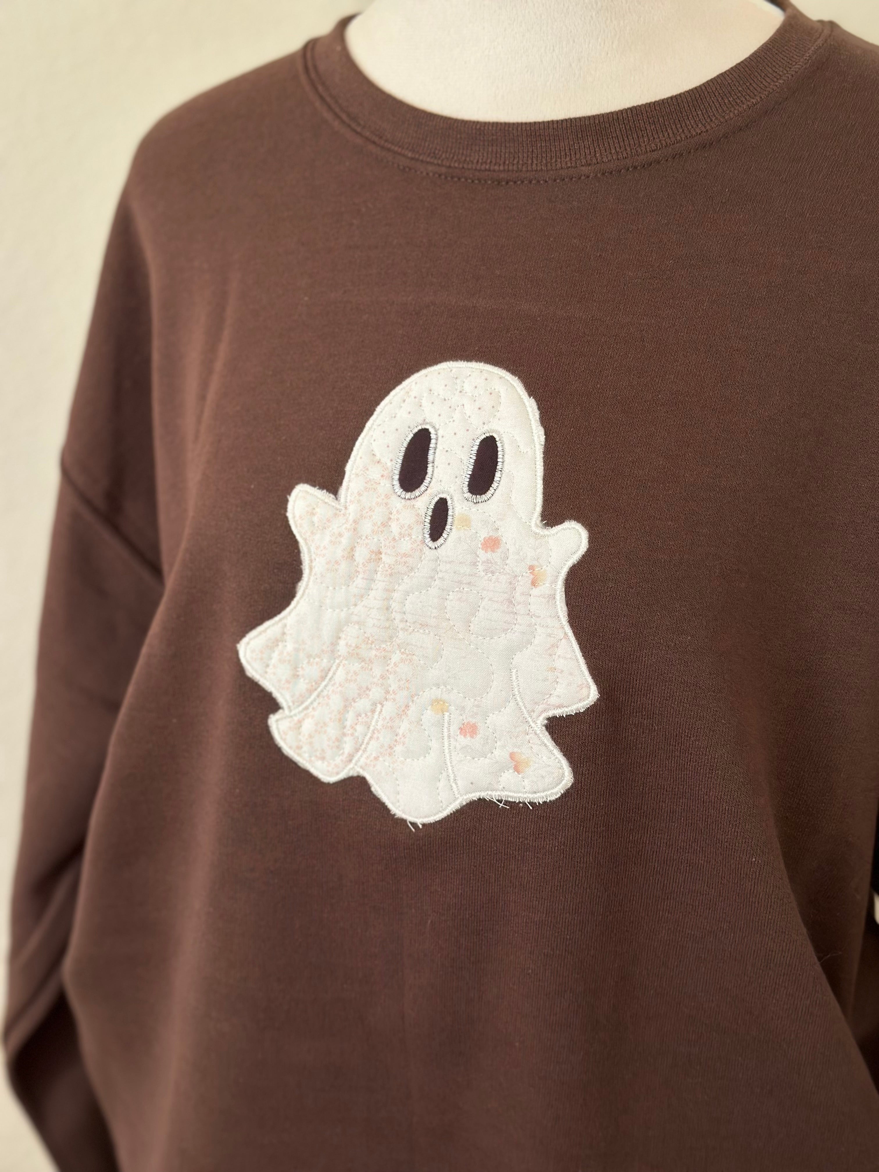 Angled view of embroidered ghost applique on the front of a brown sweatshirt.