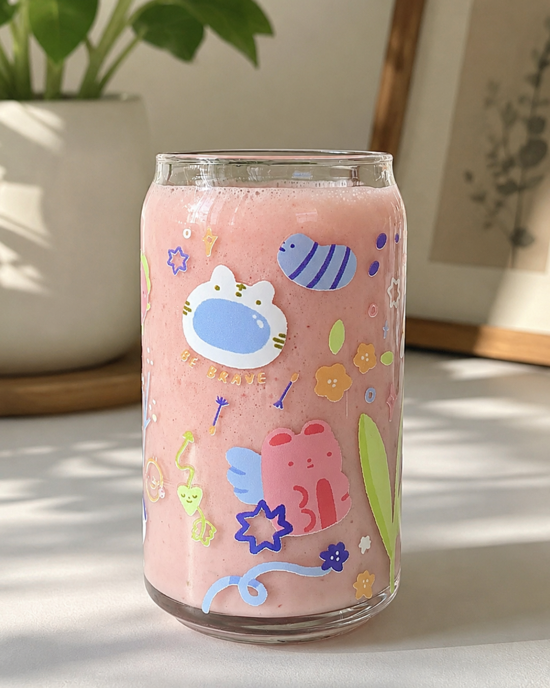 A clear glass can-shaped tumbler filled with pink smoothie, decorated with colorful cartoon stickers including a blue fish, a white cat face, a pink frog, and small flowers, set on a sunlit tabletop with a blurred plant and framed art in the background.