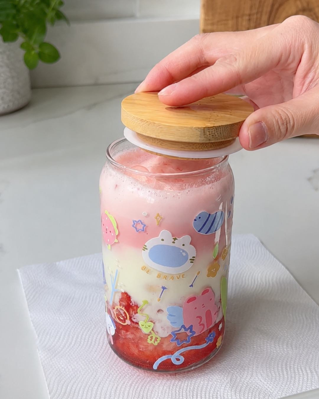 A hand is shown pressing a round wooden lid onto a clear glass jar filled with pink and red layered drink or gel, decorated with colorful cartoon characters. The jar sits on a white paper towel on a light countertop.