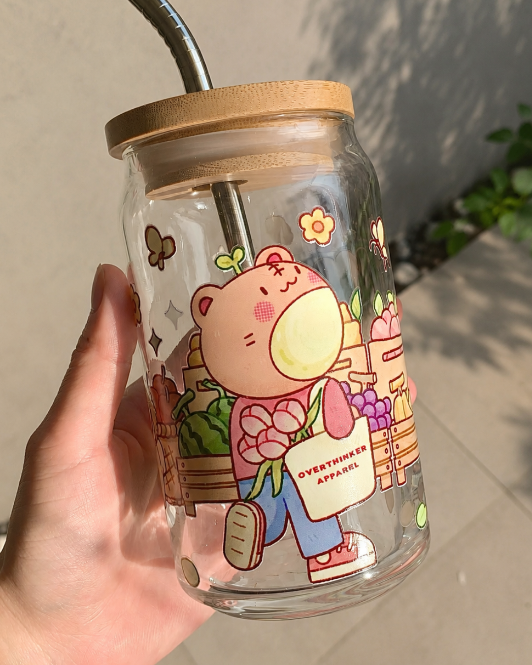 A hand holding a clear glass jar tumbler with a bamboo lid and metal straw. The tumbler features a cute cartoon illustration of a pink bear holding flowers and a bag that reads 'OVERTHINGS APPAREL'.