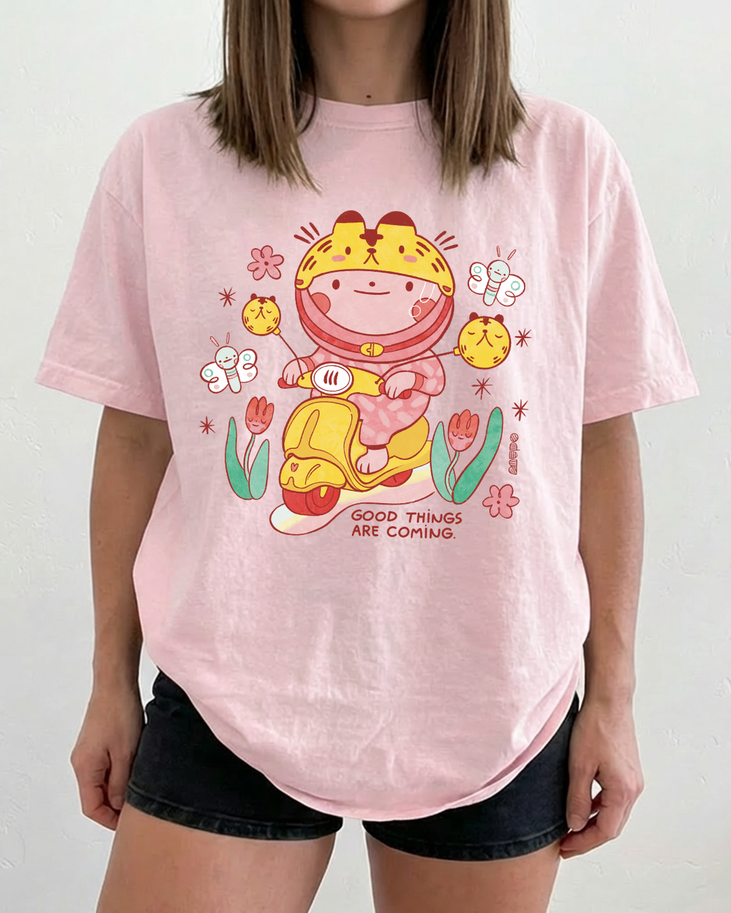 Pink graphic t-shirt featuring a cute illustrated scooter scene with flowers and the words “good things are coming”