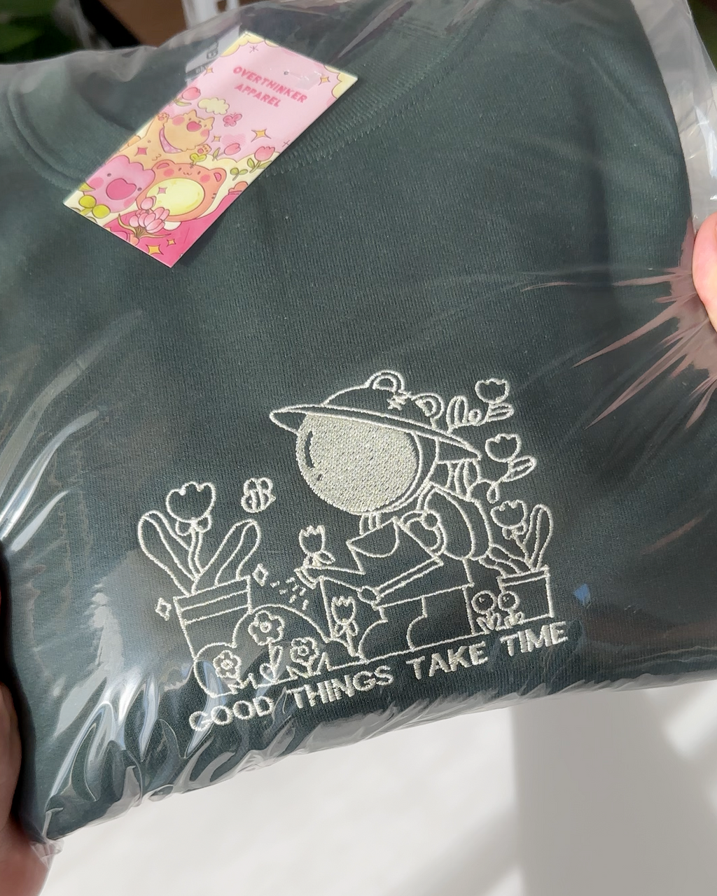 Detailed view of the embroidered gardener illustration and “Good Things Take Time” text on a forest green sweatshirt.