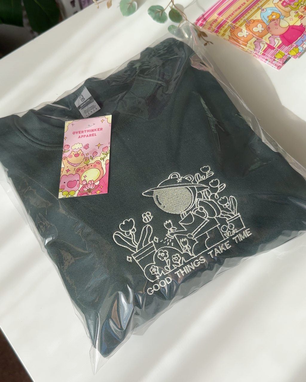 Folded forest green embroidered sweatshirt packaged with brand tag, featuring the “Good Things Take Time” gardener design.