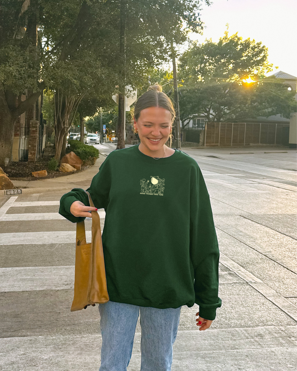 Forest green sweatshirt with ‘Good things take time’ gardening design featuring a character watering plants, worn casually outdoors with jeans and shoulder bag