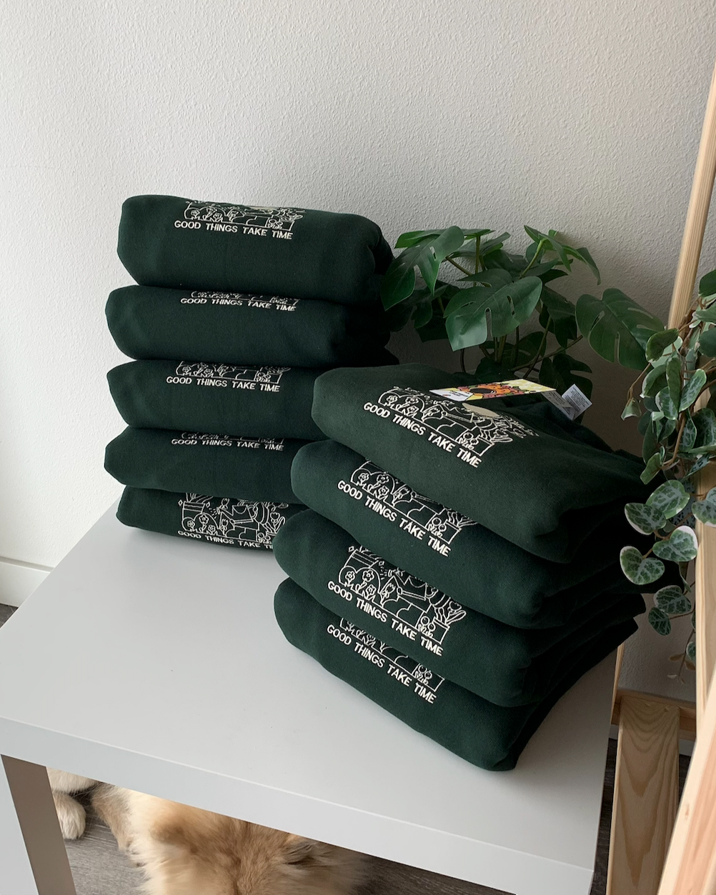Stack of dark green ‘Good things take time’ sweatshirts folded neatly on a white table with houseplants, dog peeking out underneath