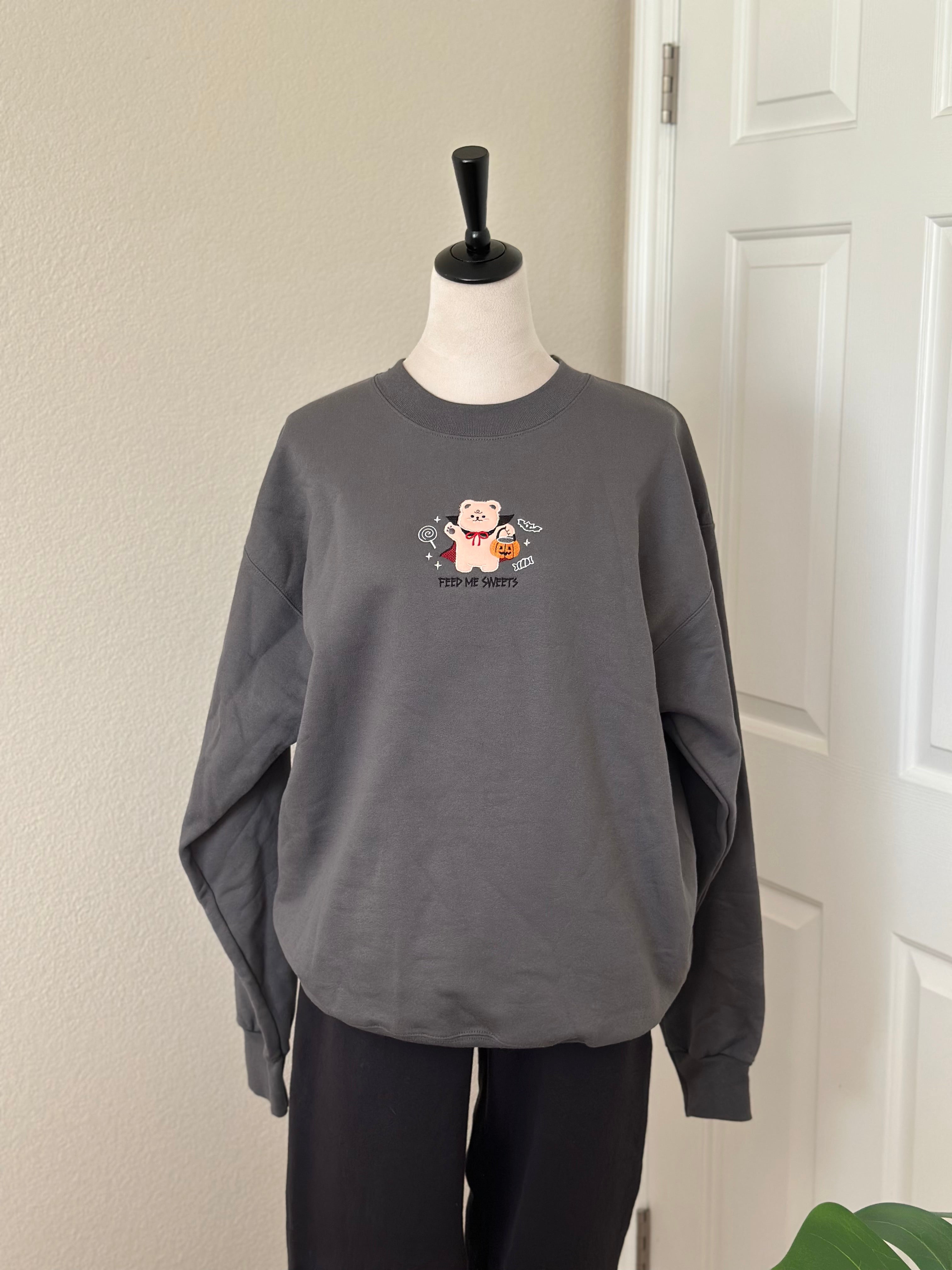 Full front view of gray sweatshirt with embroidered vampire bear, pumpkin bucket, and candy with “Feed Me Sweets” text.