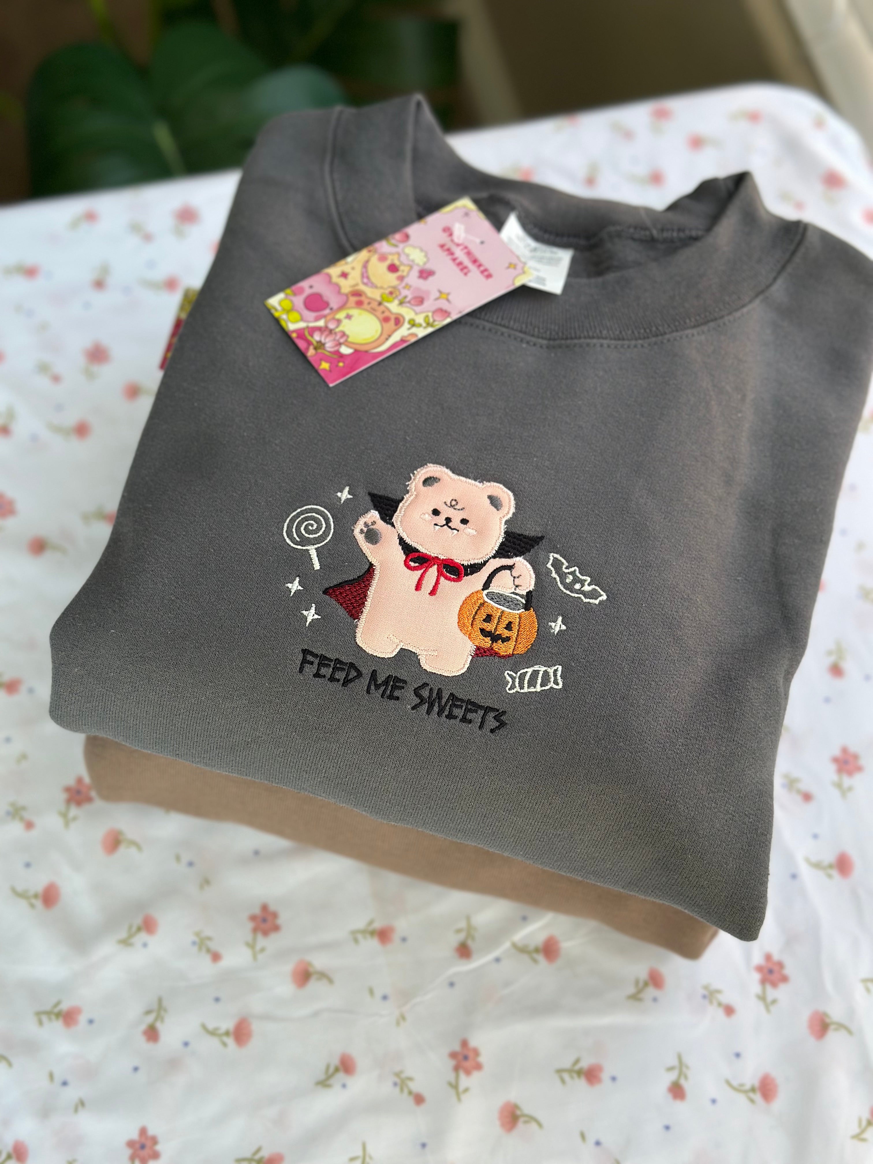 Folded gray sweatshirt with embroidered vampire bear holding pumpkin candy bucket and lollipop, “Feed Me Sweets” text visible.