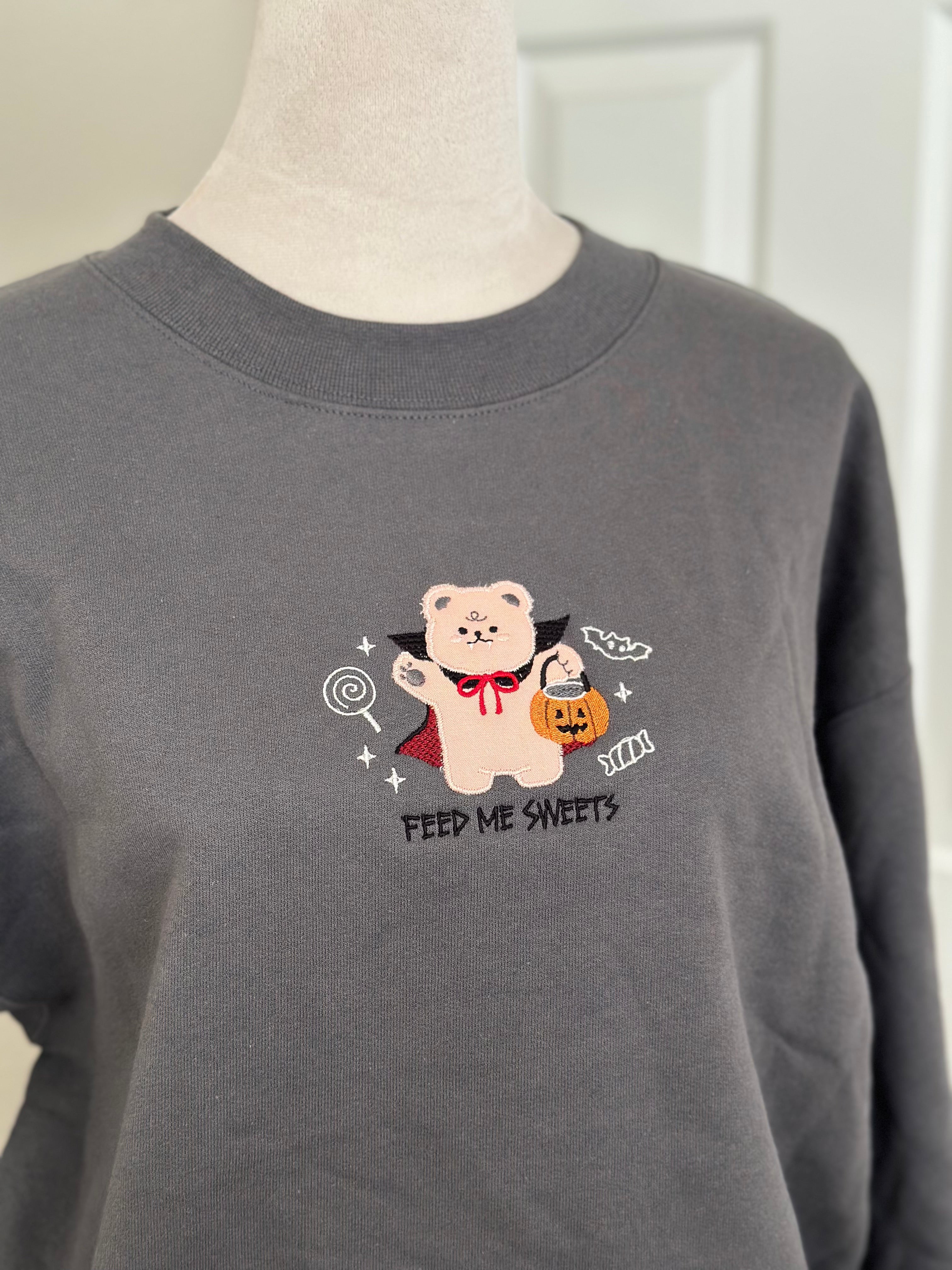 Close-up of a gray sweatshirt with an embroidered bear in a vampire cape holding a pumpkin bucket, surrounded by candy and the words “Feed Me Sweets.”