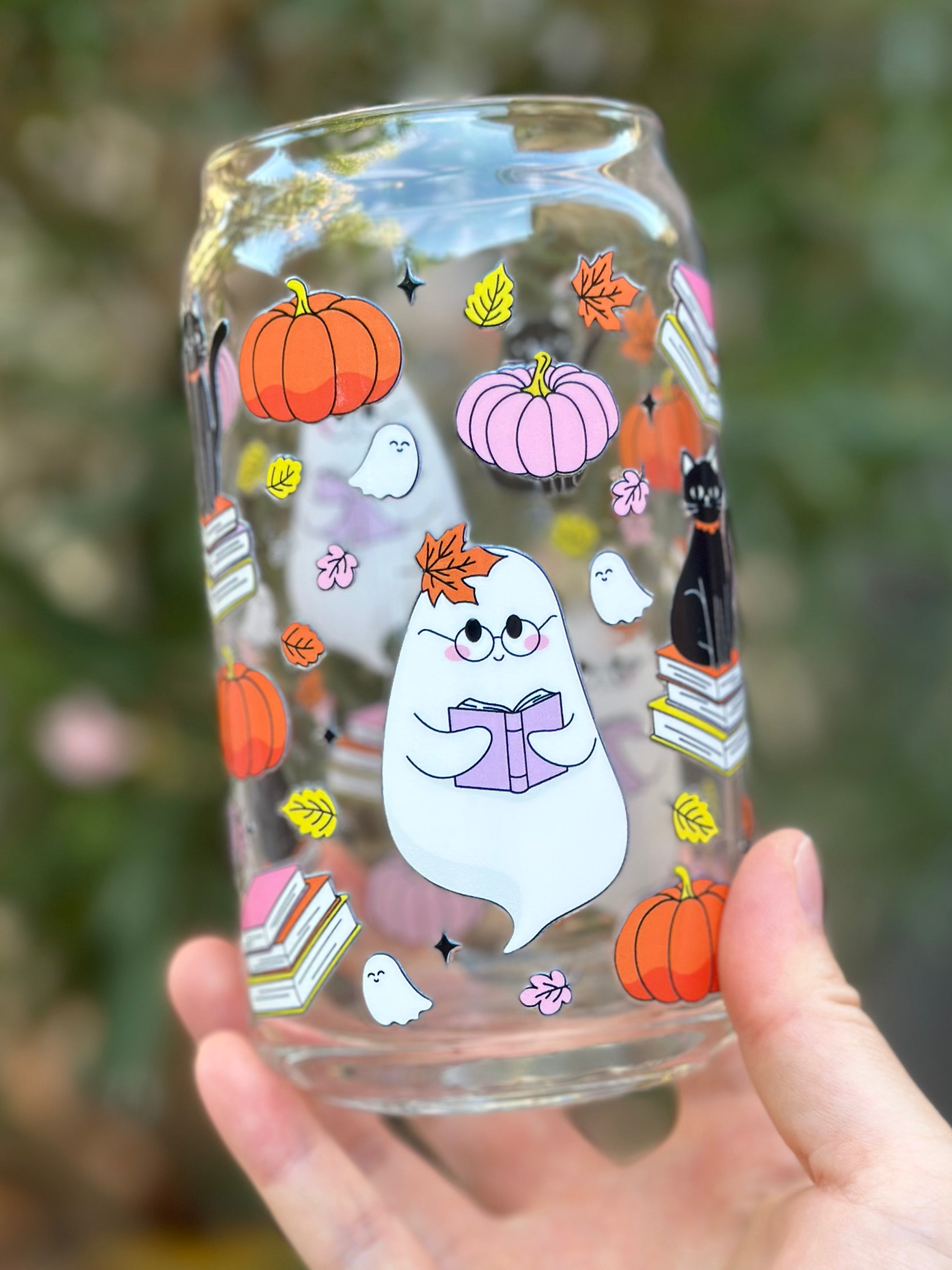 Glass can cup with a reading ghost, pumpkins, pink books, black cat, and colorful autumn leaves.