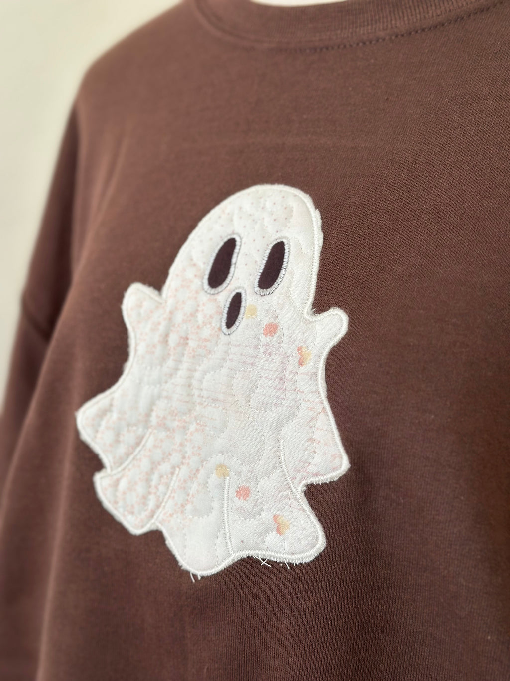 Angled close-up of embroidered white ghost with stitched details on brown sweatshirt.