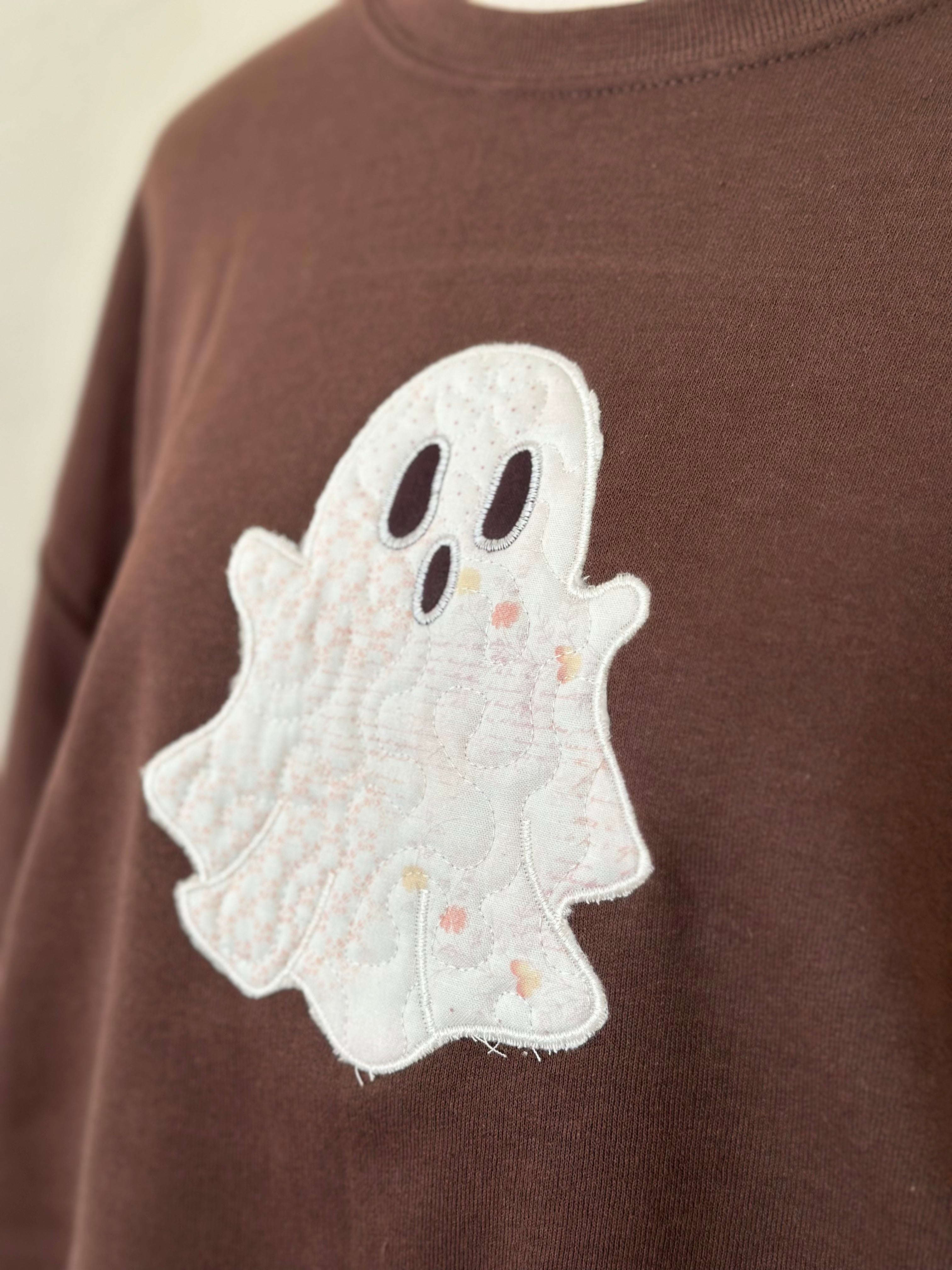 Angled close-up of embroidered white ghost with stitched details on brown sweatshirt.