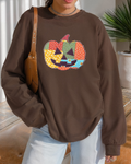 Person standing indoors wearing a brown sweatshirt with patchwork jack-o’-lantern, paired with jeans and shoulder bag.