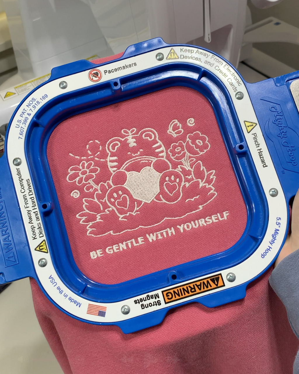 Hand embroidery process showing a self compassion message stitched onto a rose pink crewneck sweatshirt