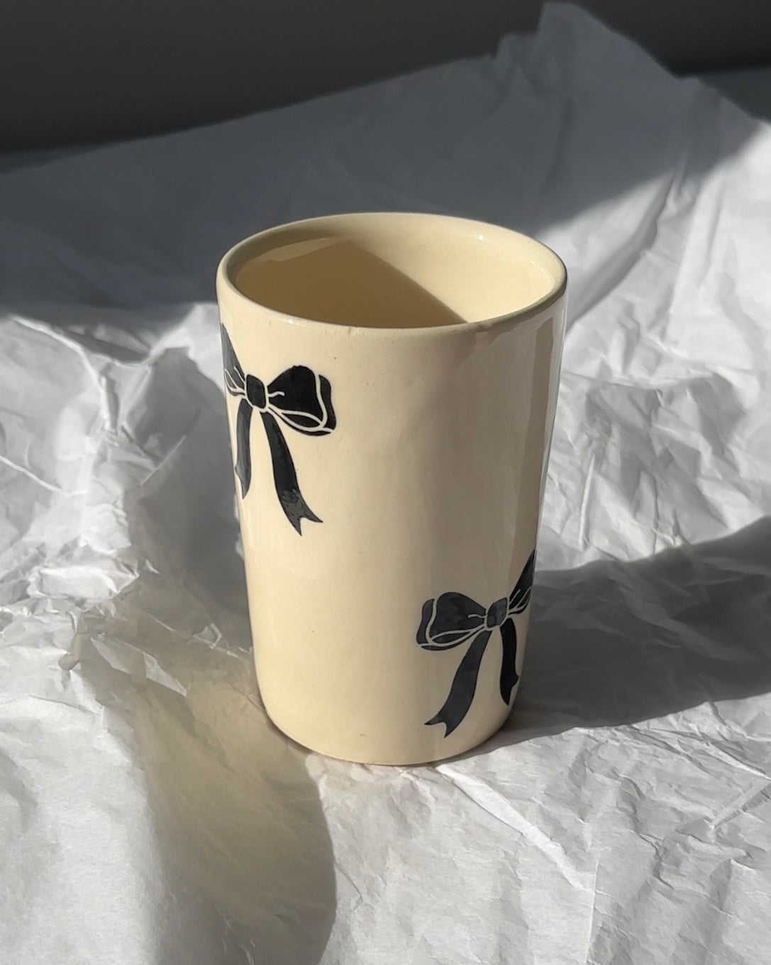 Hand-painted stoneware mug with black bow details on cream glaze, handmade ceramic coffee cup in natural light
