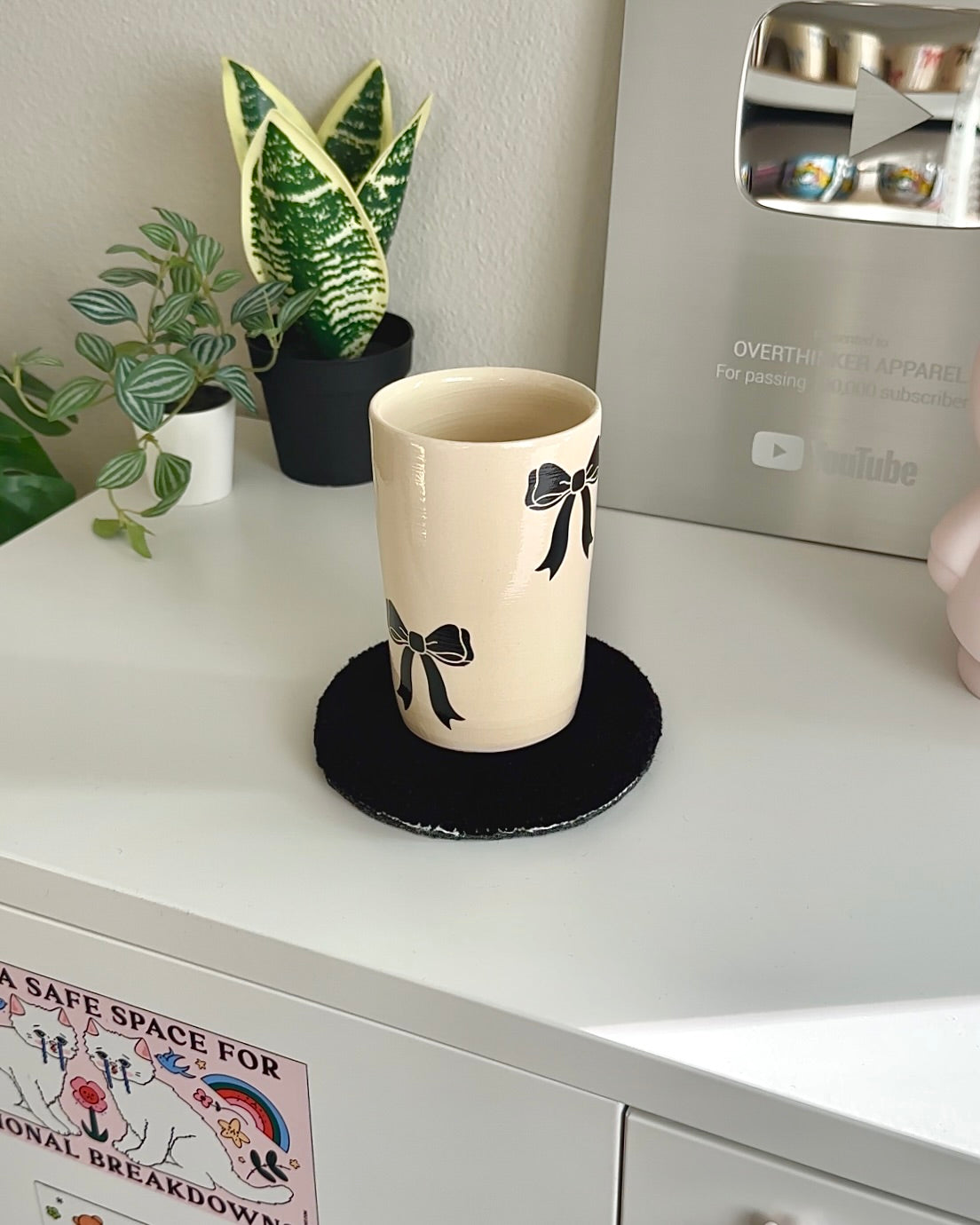 Handmade cream stoneware coffee mug with black bow design, styled on a desk with plants for a neutral coquette aesthetic

