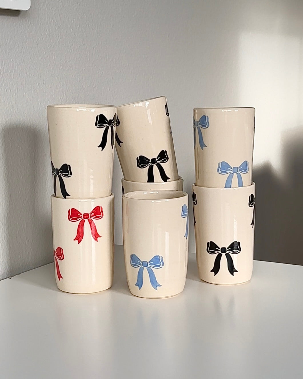 Handmade stoneware ceramic mugs with bow designs in blue, black, and red, displayed together as a small-batch pottery collection
