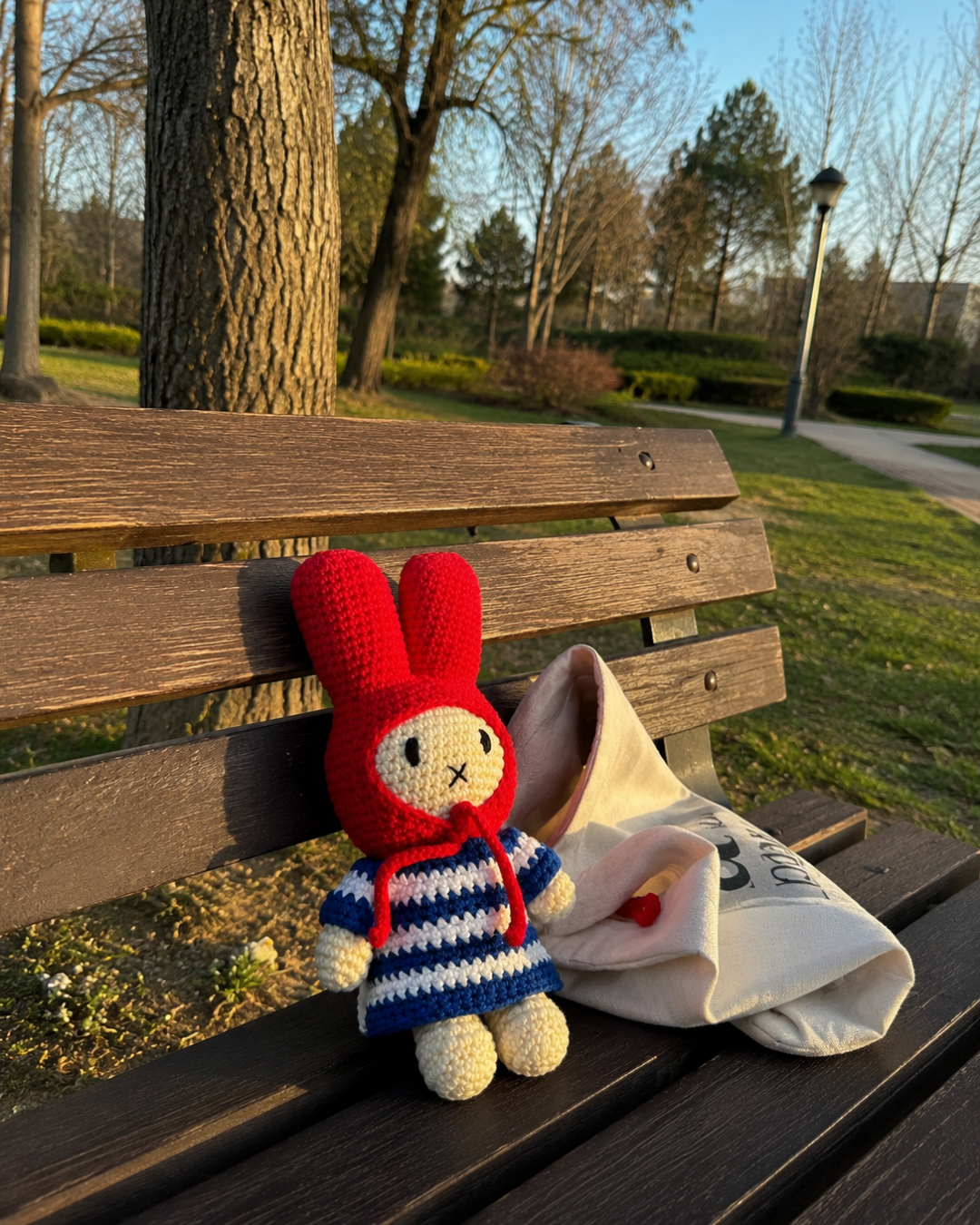 A red crocheted bunny doll wearing a blue and white striped sweater sits on a wooden park bench next to a cream tote bag, with trees and grass in the background.