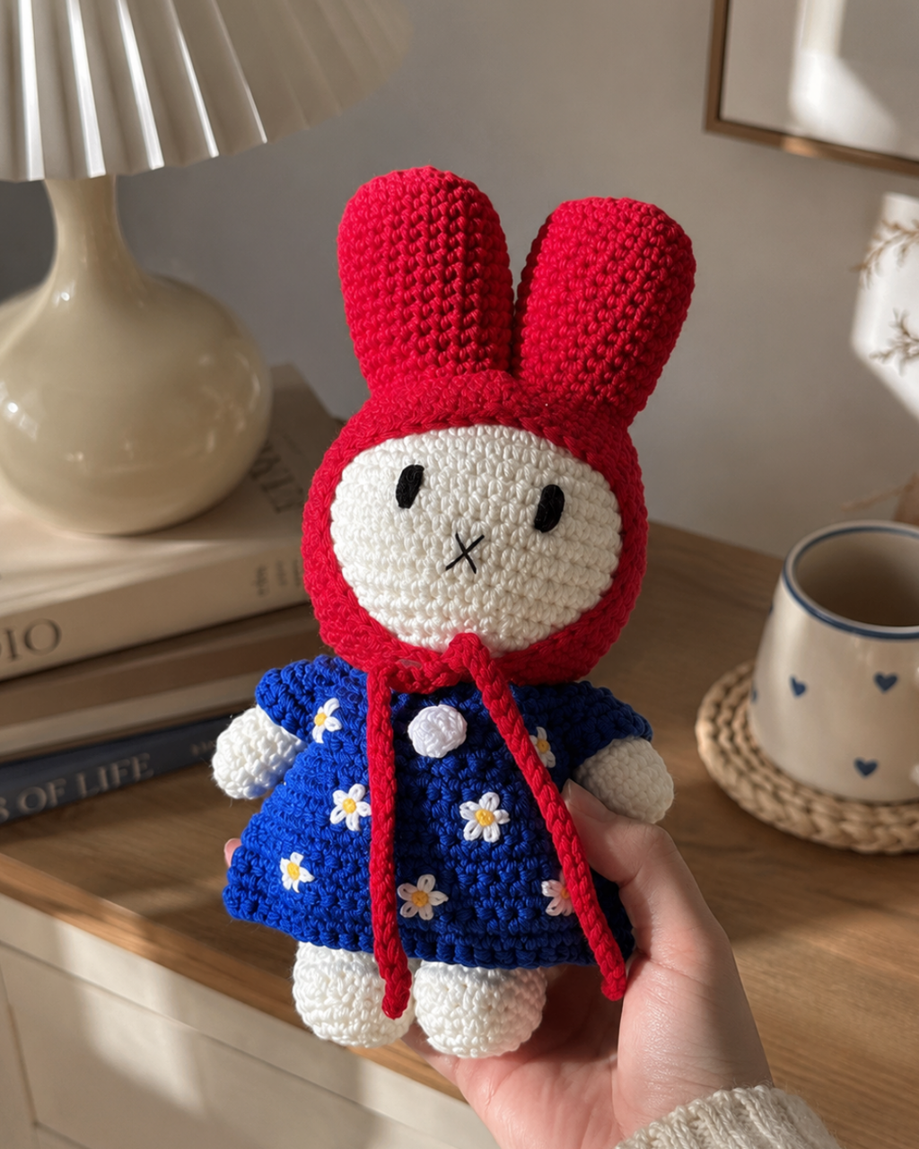 A crocheted rabbit doll with a red bonnet and blue dress decorated with white flowers, held in a hand over a wooden table with a lamp and mug in the background.