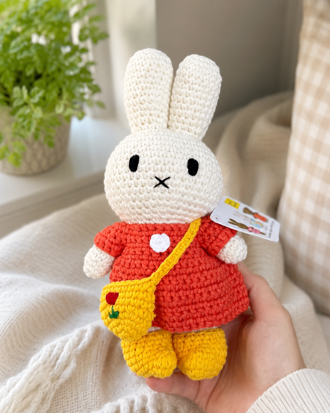 A crocheted plush bunny doll wearing a red dress and yellow shoes, held in a person's hand against a soft indoor background with a plant and cream-colored textiles.