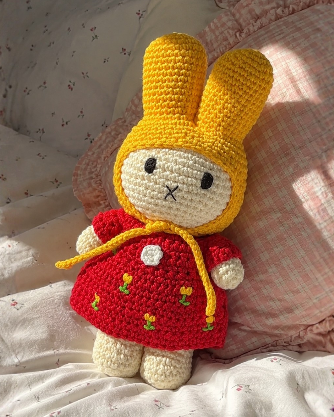 A crocheted bunny doll wearing a red dress with small yellow and green flower details, posed on a bed with pink and white patterned bedding.