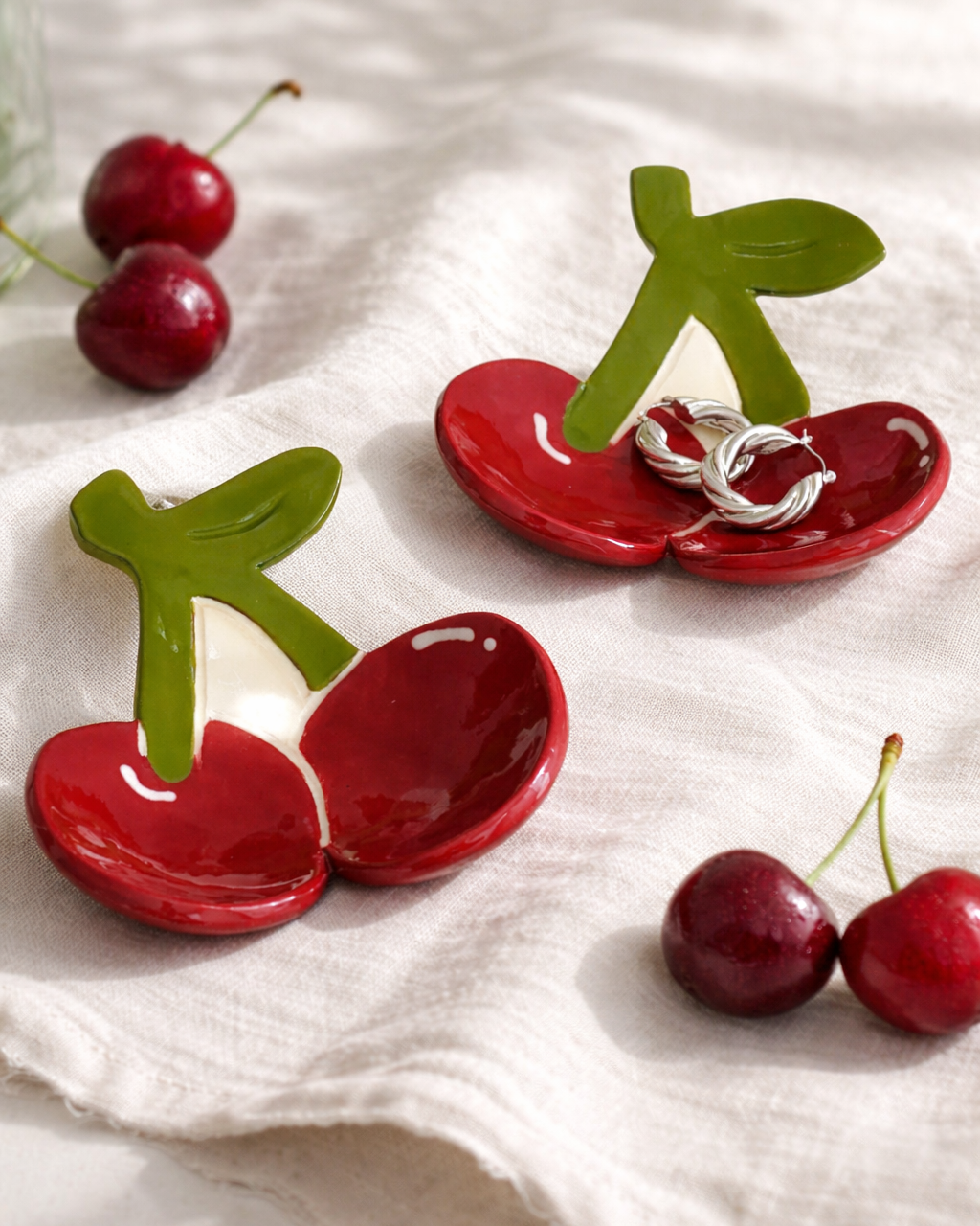 Handmade ceramic cherry jewelry dish holding hoop earrings on linen cloth with fresh cherries, fruit shaped trinket dish for vanity decor and jewelry storage