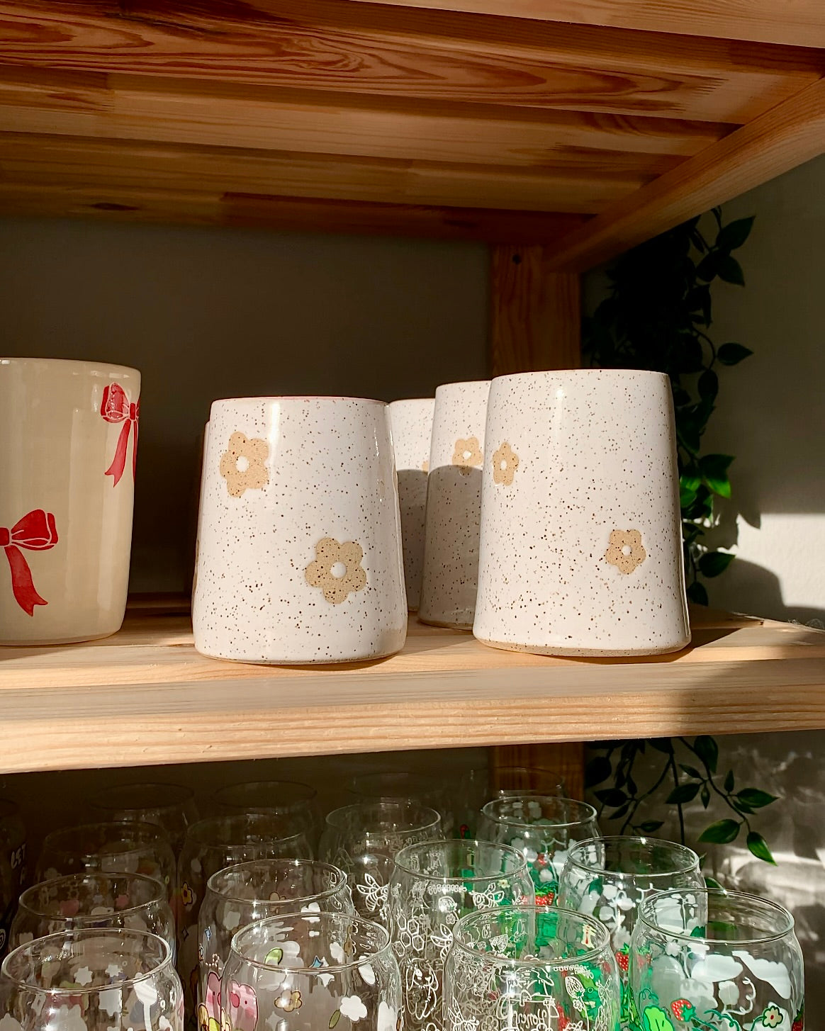 Handmade speckled stoneware daisy mugs displayed on a wooden shelf, showcasing small-batch ceramic pottery in warm light
