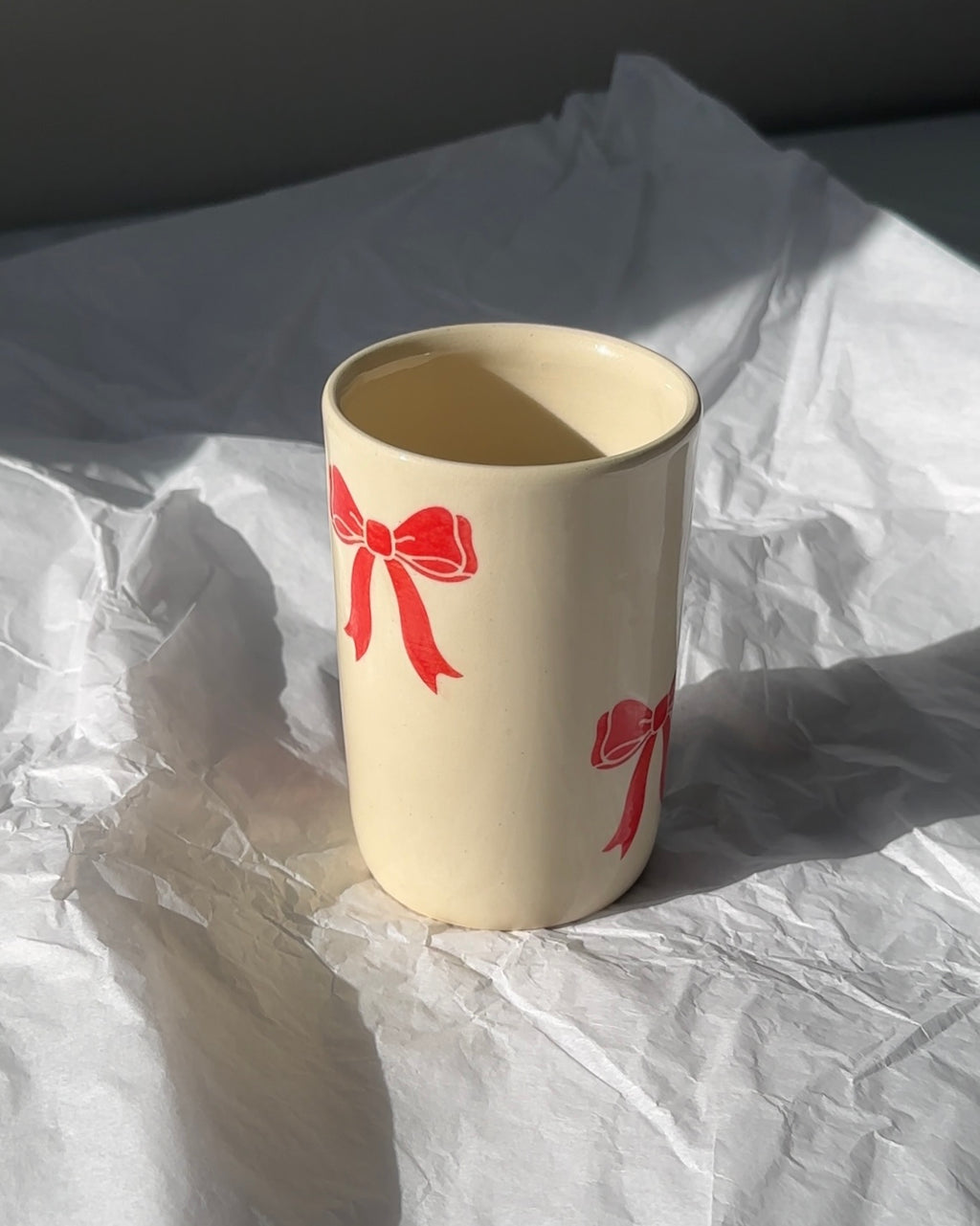 Handmade ceramic mug with hand-painted red bow design on a cream stoneware glaze, shown upright in natural light, coquette-inspired artisan coffee cup.