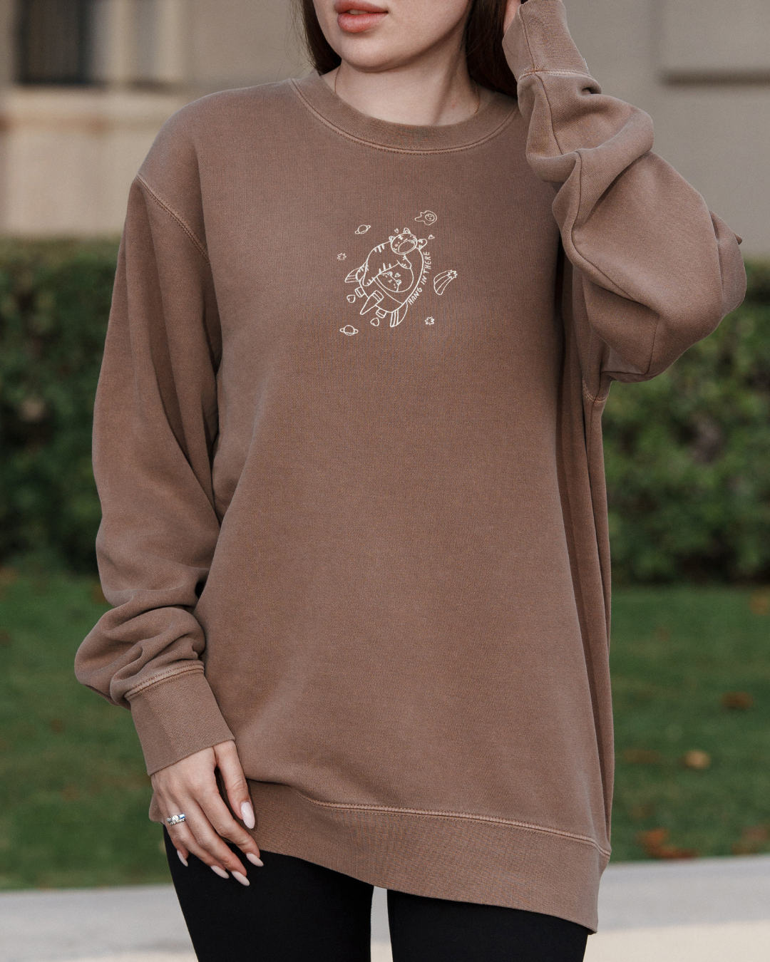 Model wearing a mocha brown embroidered crewneck sweatshirt featuring a small floating character and the words “Hang In There,” a subtle emotional support sweatshirt designed for comfort, burnout days, and everyday wear.

