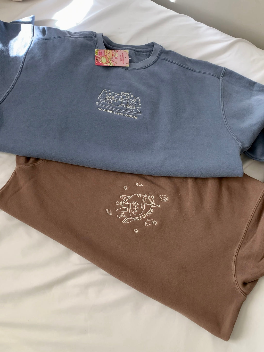Mocha brown embroidered sweatshirt stacked on a blue crewneck, showing the subtle “hang in there” character design on the chest.