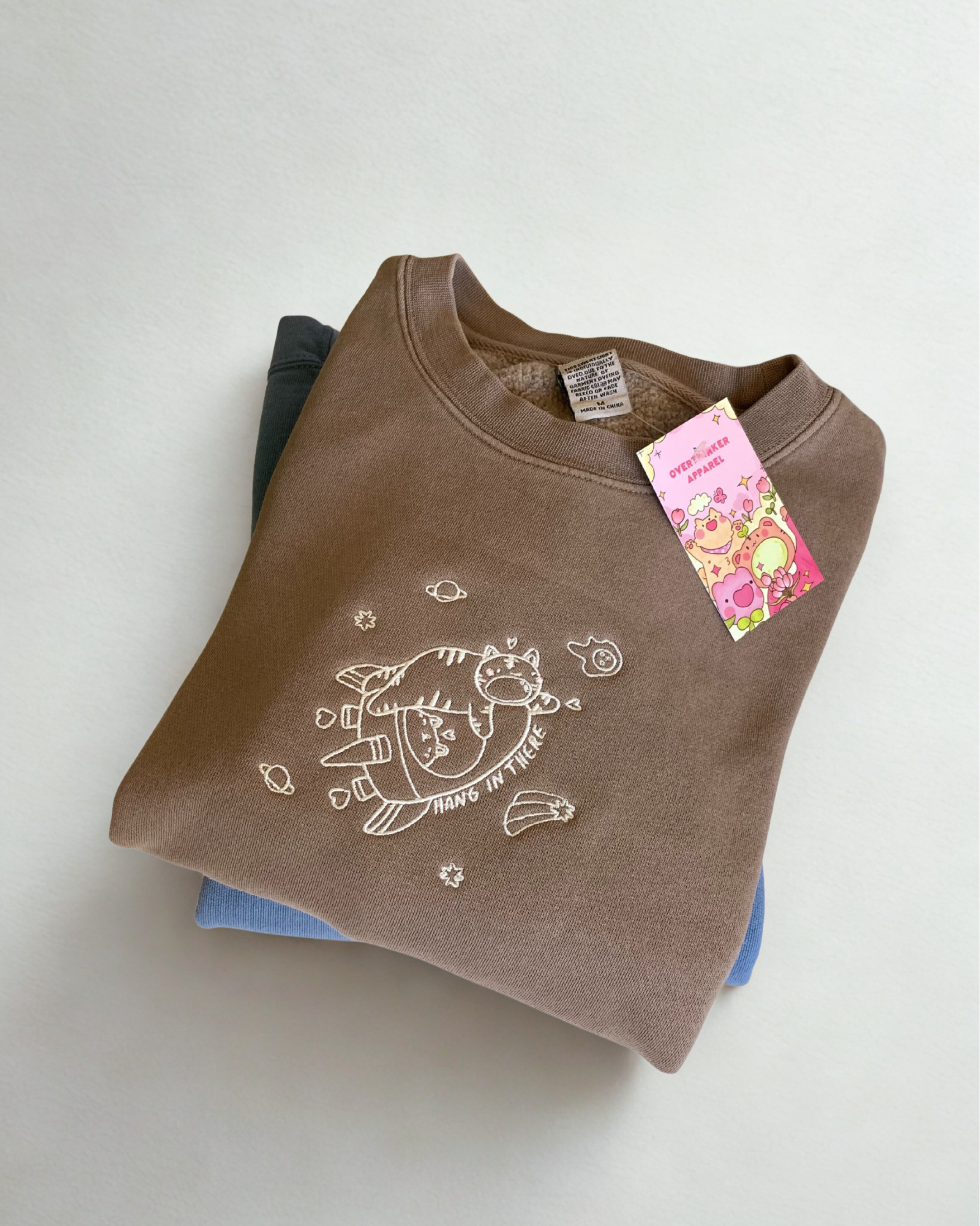Close-up of embroidered “hang in there” design on a mocha brown sweatshirt, showing stitching detail and soft fleece fabric.