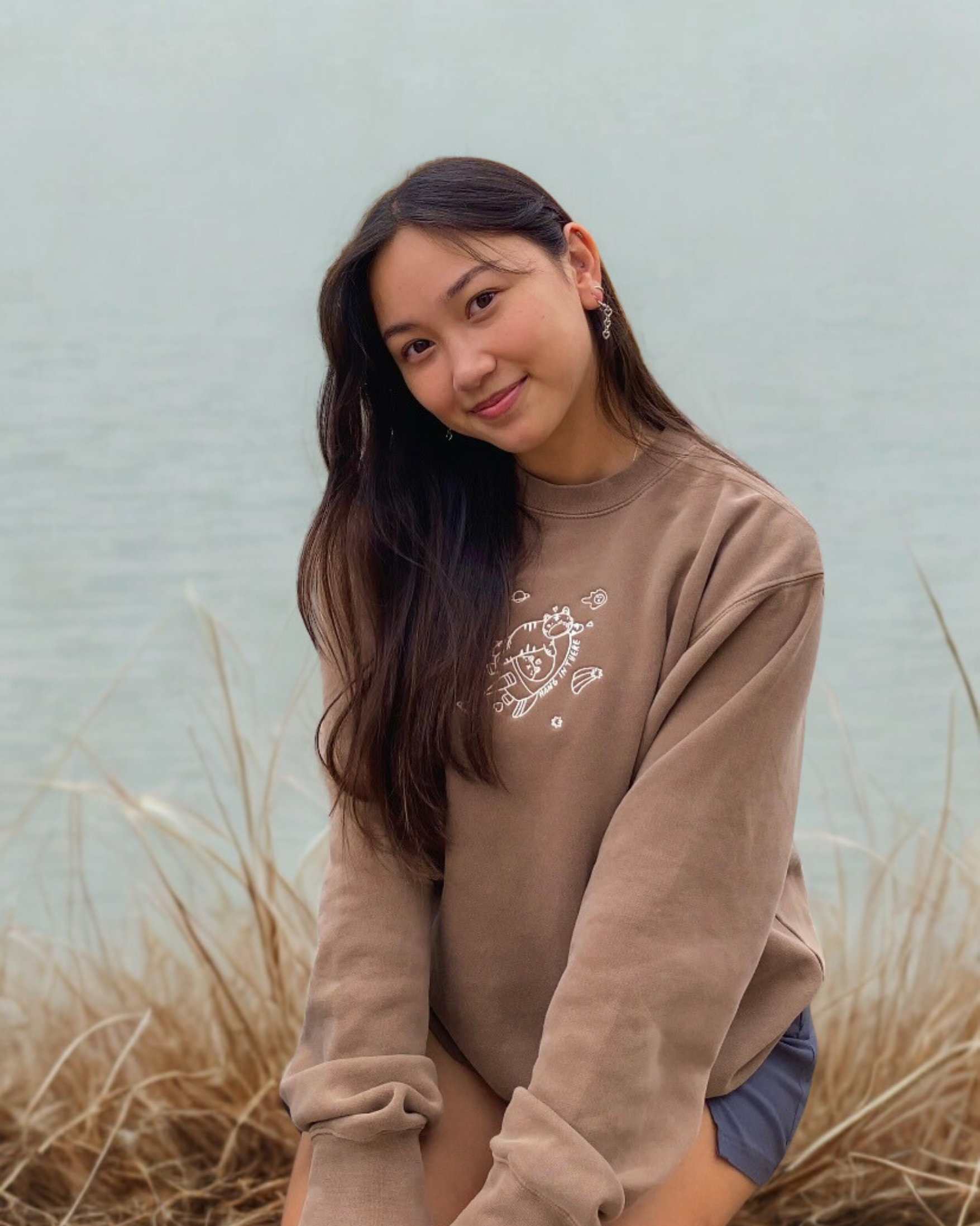 Model wearing a mocha brown embroidered sweatshirt with a small “hang in there” character design, seated near the water.