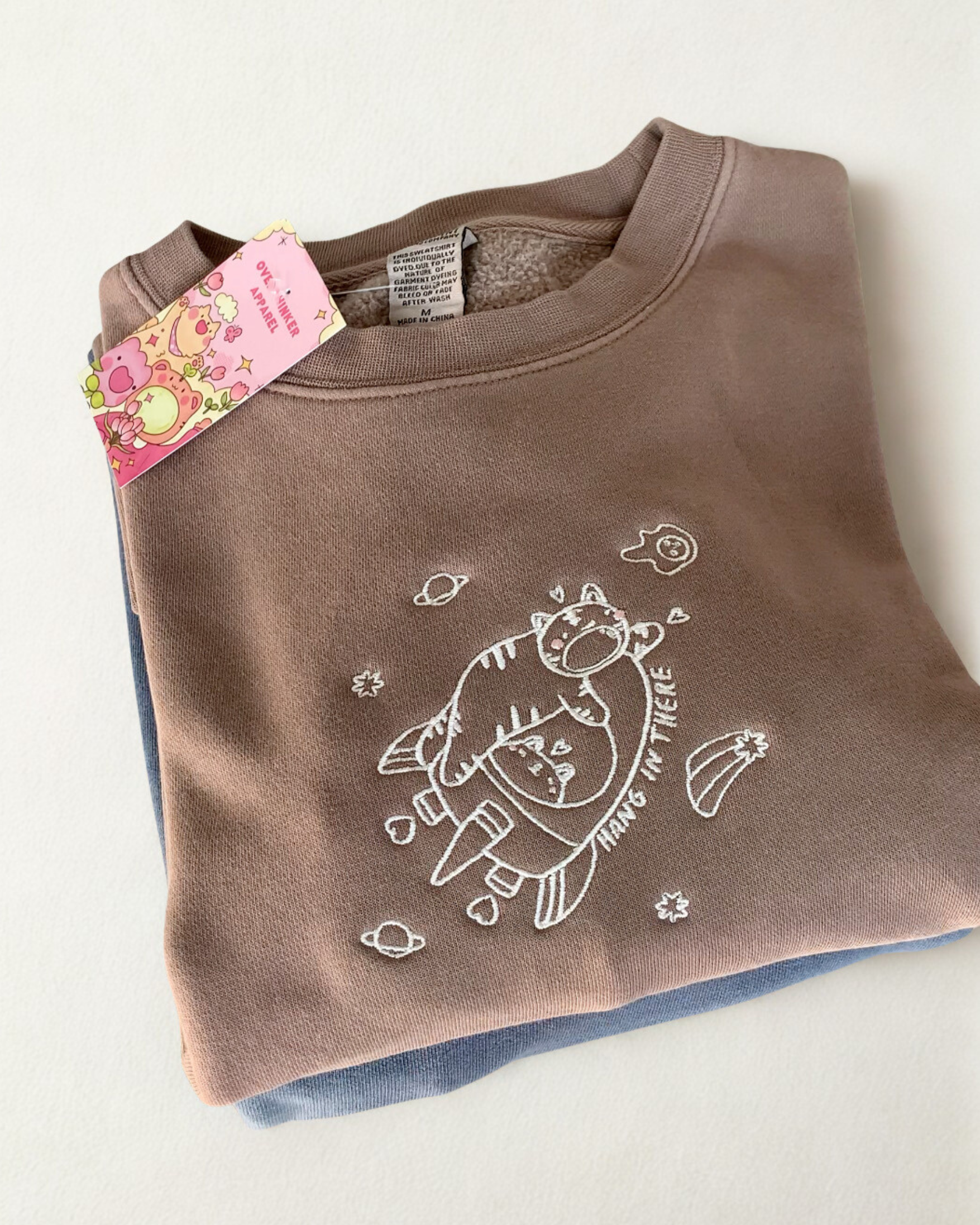 Folded mocha brown crewneck sweatshirt featuring a small embroidered character floating among stars with the words “hang in there.”
