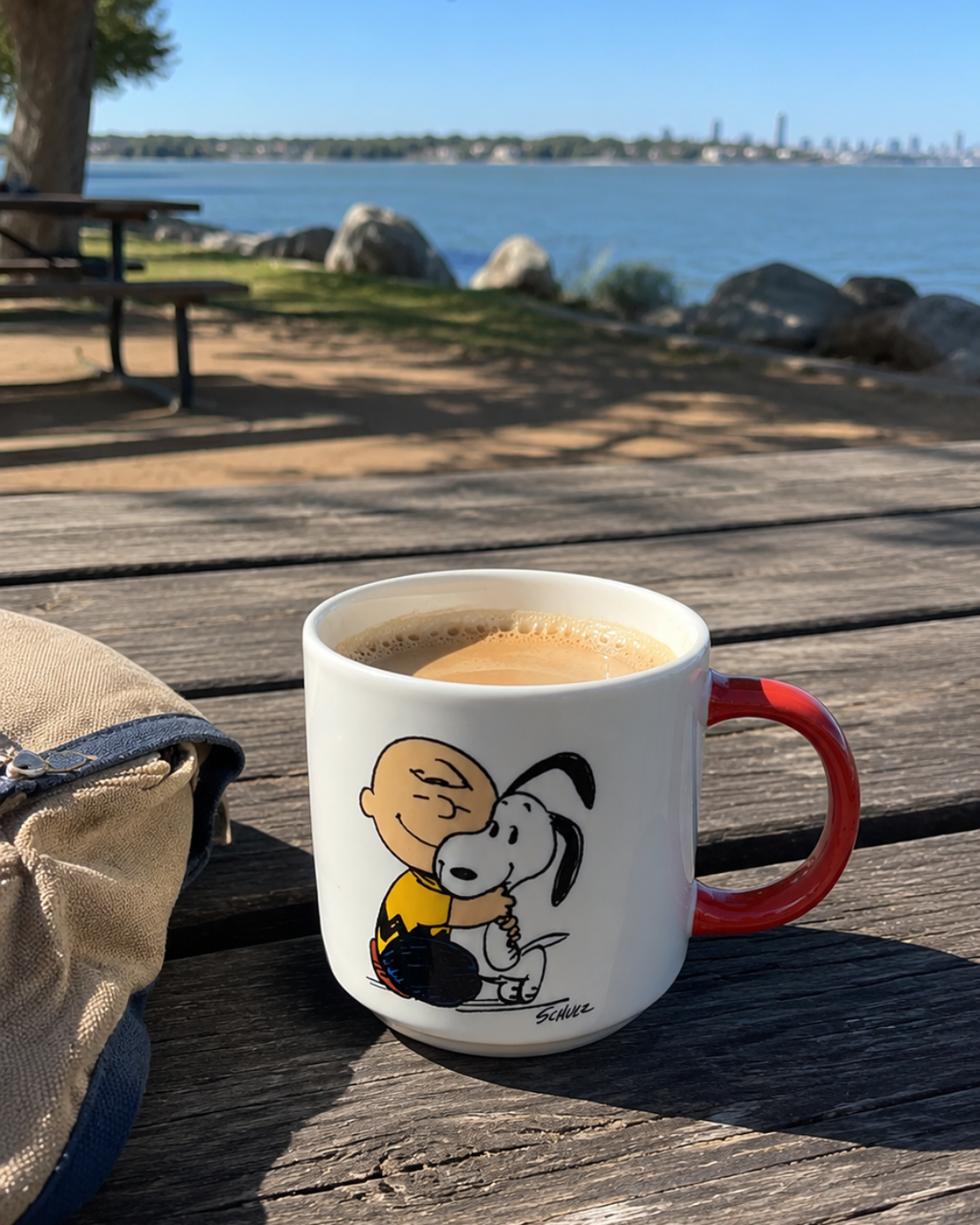 A Snoopy mug with Charlie Brown and Snoopy sits on a wooden picnic table by the water, with a city skyline in the background, filled with coffee.