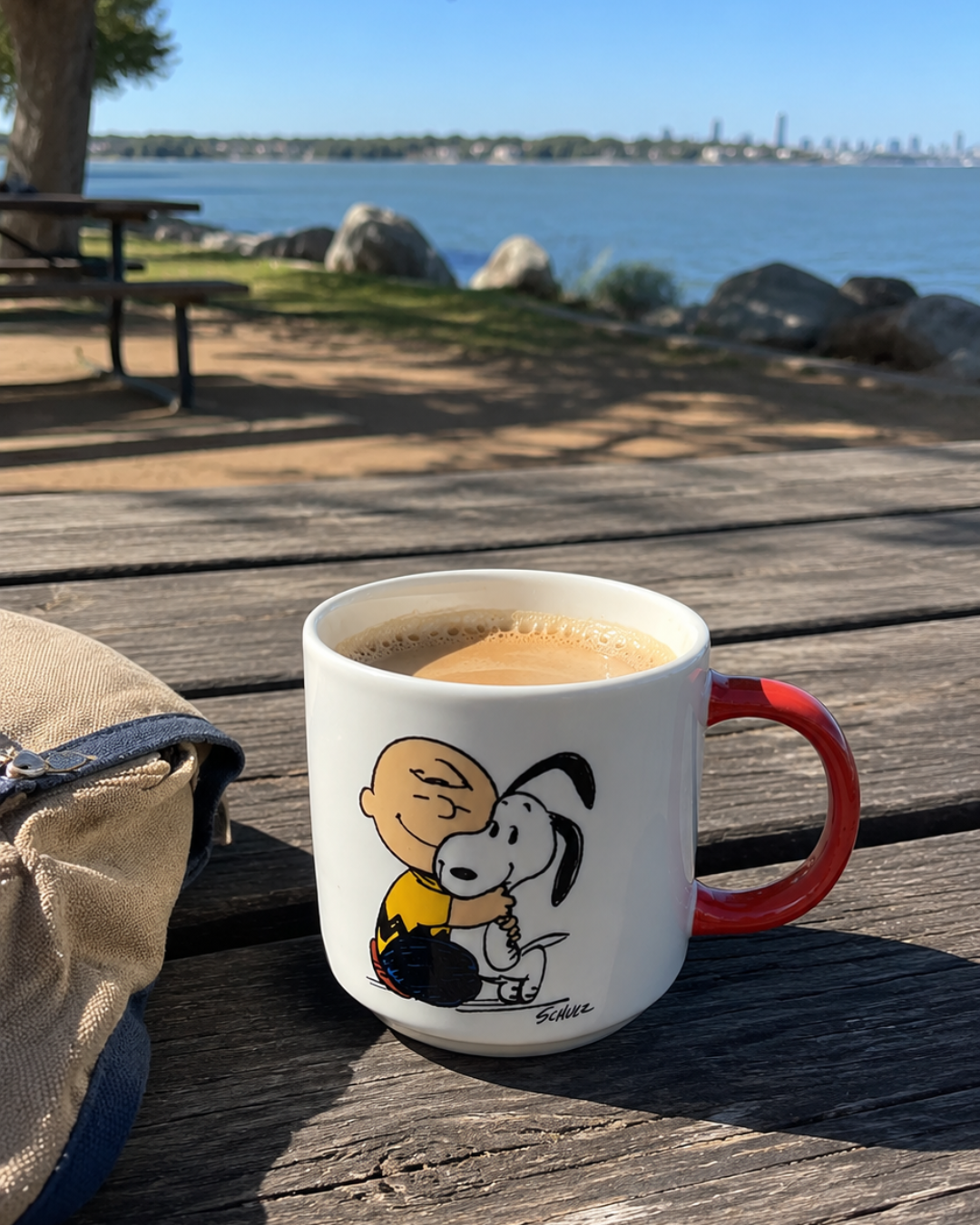 A Snoopy mug with Charlie Brown and Snoopy sits on a wooden picnic table by the water, with a city skyline in the background, filled with coffee.
