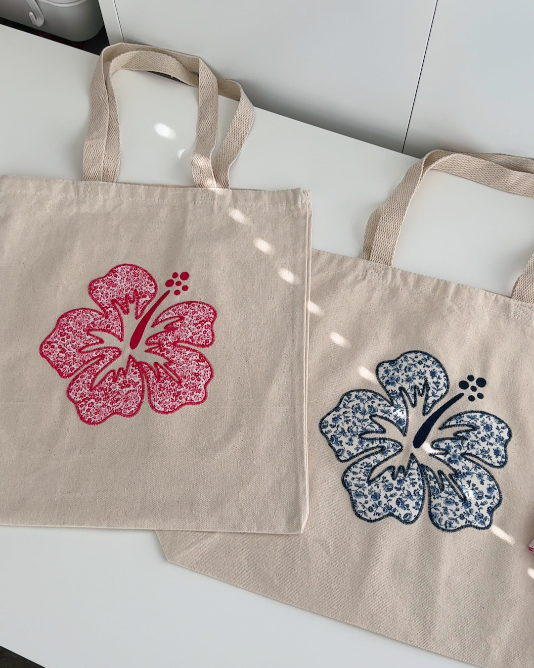 Natural canvas tote bags with pink and navy hibiscus embroidery shown flat lay, floral patchwork design in regular and oversized sizes