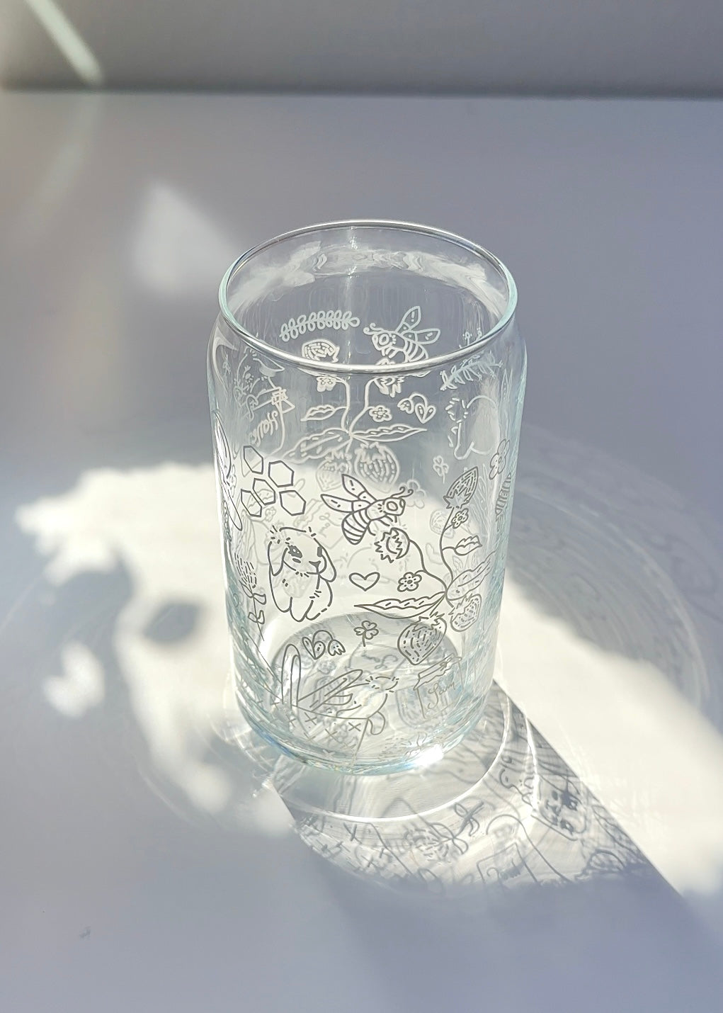 Clear 16 oz glass can with a cottagecore honey bunny illustration featuring bunnies, bees, flowers, and strawberries, styled in soft natural sunlight.