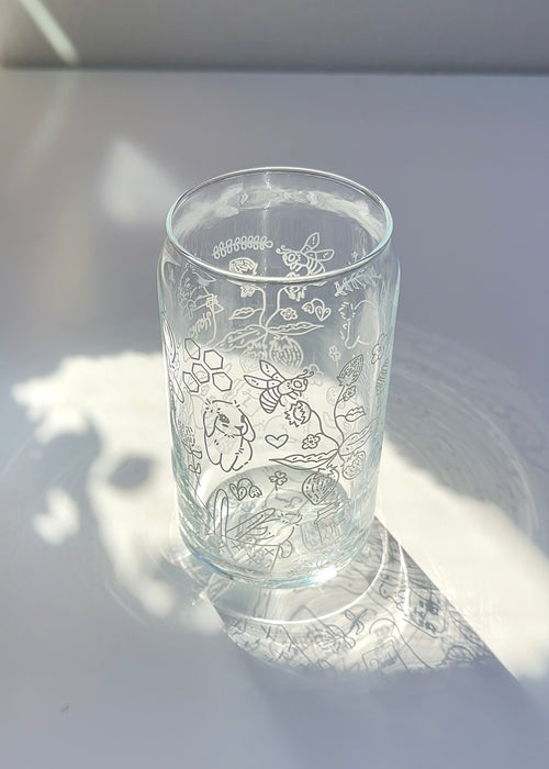 Clear 16 oz glass can with a cottagecore honey bunny illustration featuring bunnies, bees, flowers, and strawberries, styled in soft natural sunlight.