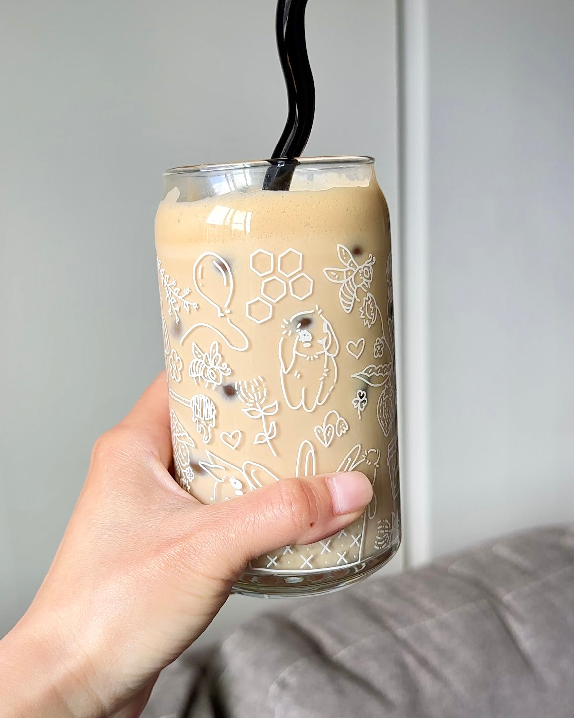 Hand holding a 16 oz honey bunny glass can filled with iced coffee, featuring white cottagecore illustrations and a reusable straw.