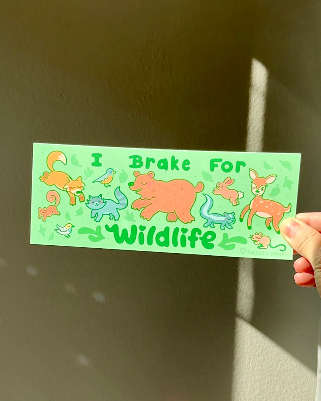 Hand holding an “I Brake for Wildlife” vinyl bumper sticker with illustrated forest animals, gentle animal lover car decal in soft green tones.