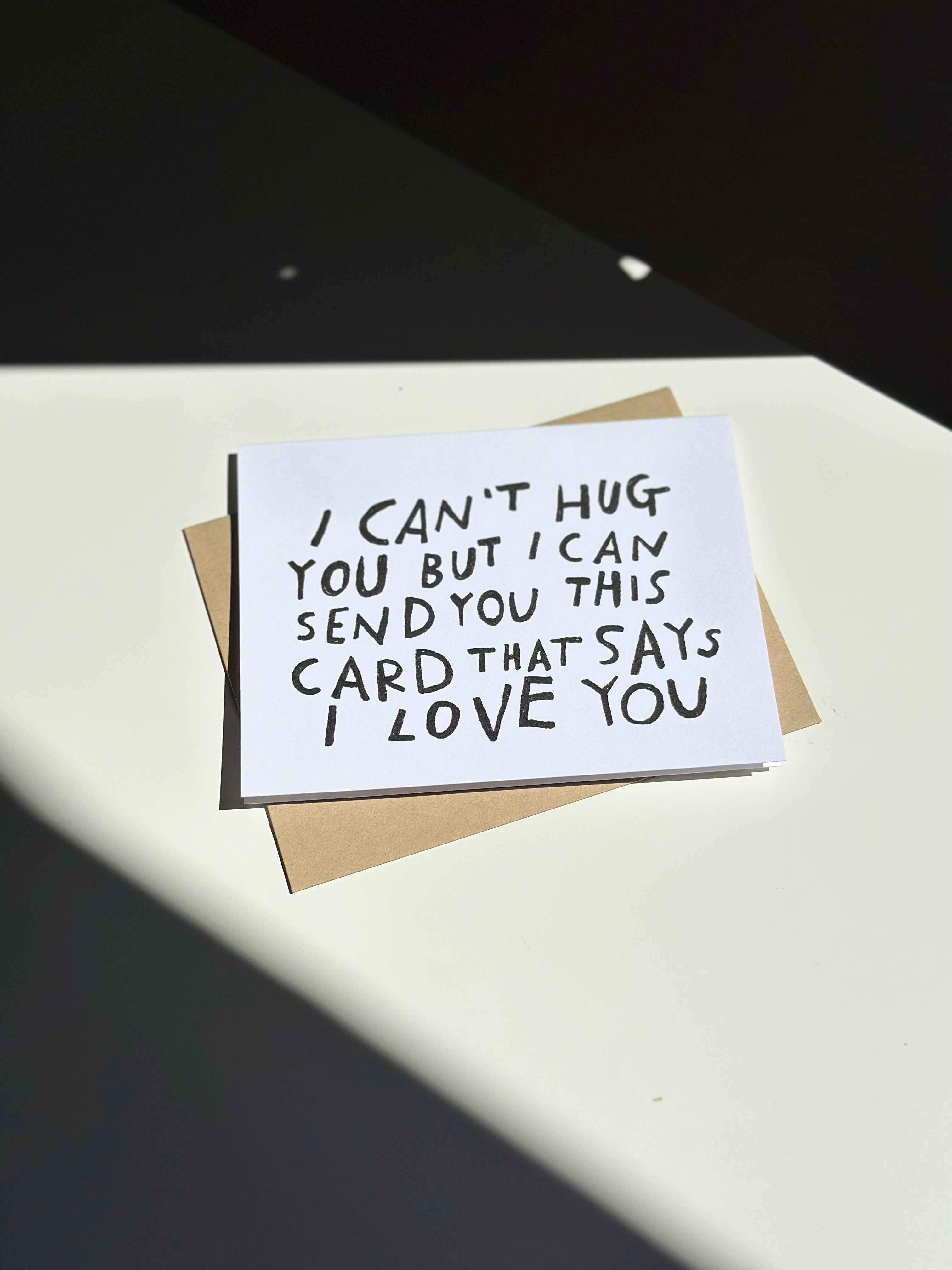 Minimalist white greeting card with black hand-lettered text that reads ‘I can’t hug you but I can send you this card that says I love you,’ paired with brown kraft envelope.