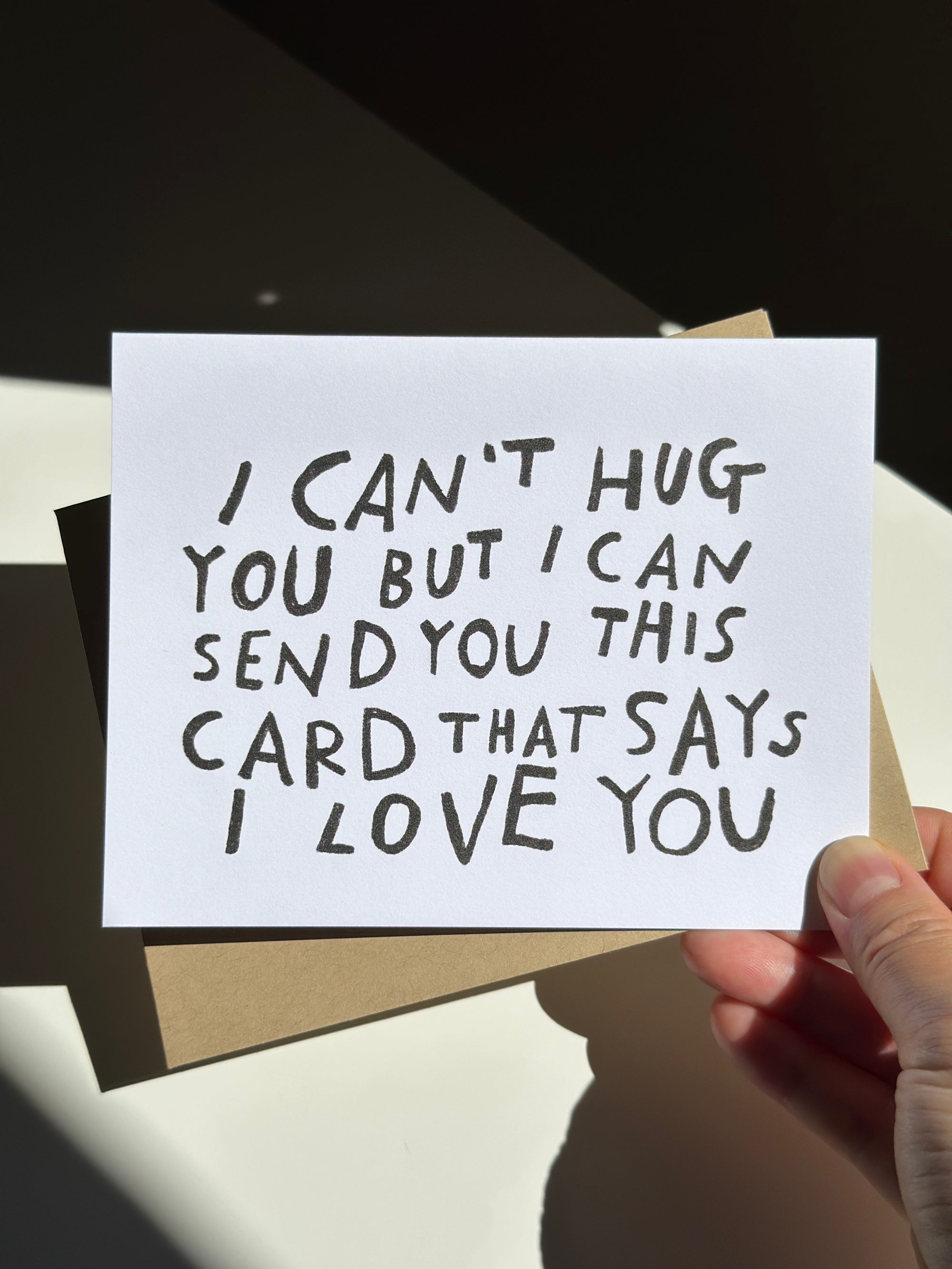 Minimalist white greeting card with black hand-lettered text that reads ‘I can’t hug you but I can send you this card that says I love you,’ paired with brown kraft envelope.