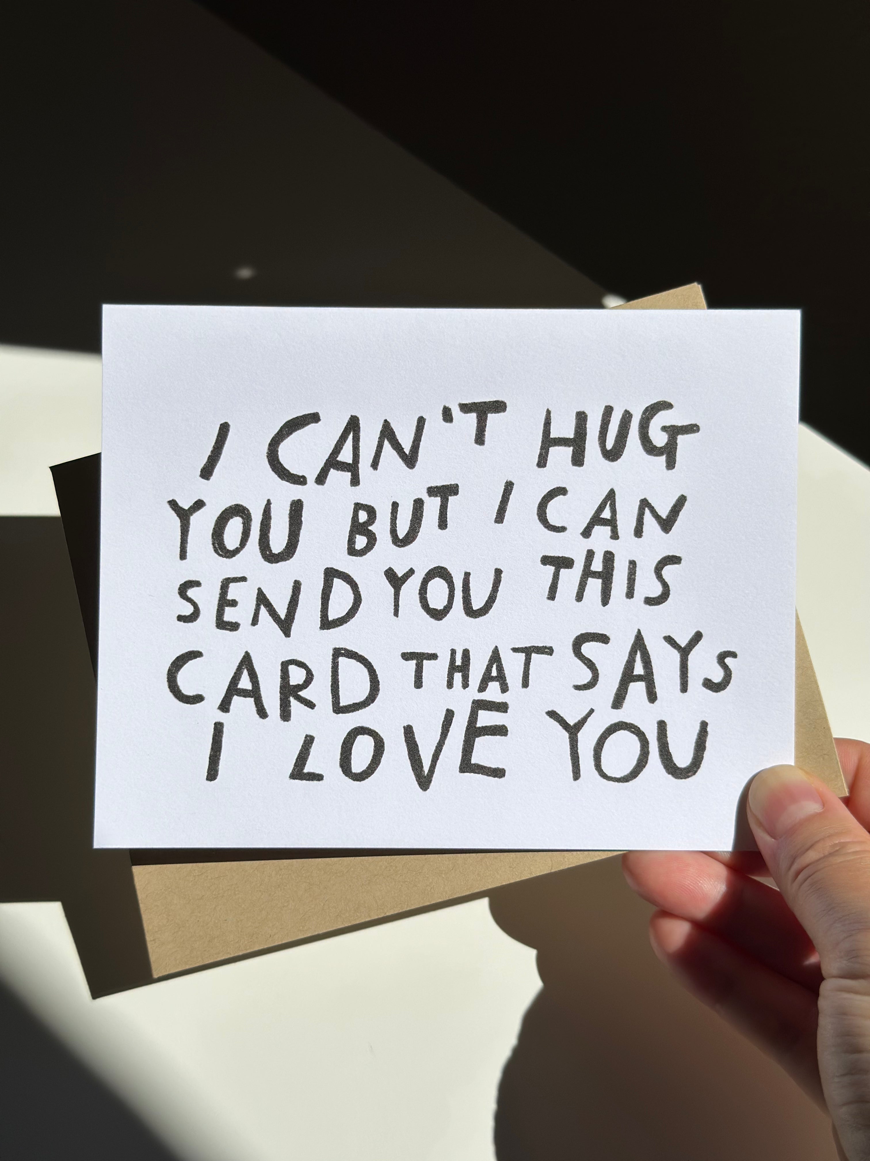 Minimalist white greeting card with black hand-lettered text that reads ‘I can’t hug you but I can send you this card that says I love you,’ paired with brown kraft envelope.