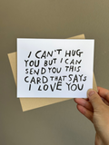 Minimalist white greeting card with black hand-lettered text that reads ‘I can’t hug you but I can send you this card that says I love you,’ paired with brown kraft envelope.