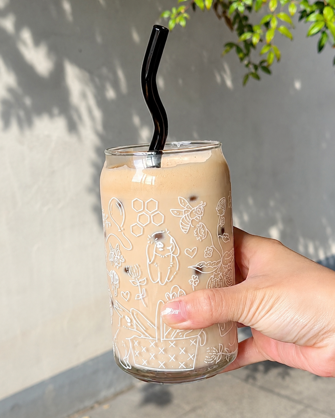 A hand holding a clear glass can-shaped tumbler filled with iced coffee, decorated with white line-art illustrations of bees, flowers, and hearts. The glass has a black curved straw and is photographed outdoors against a gray wall with leaf shadows.