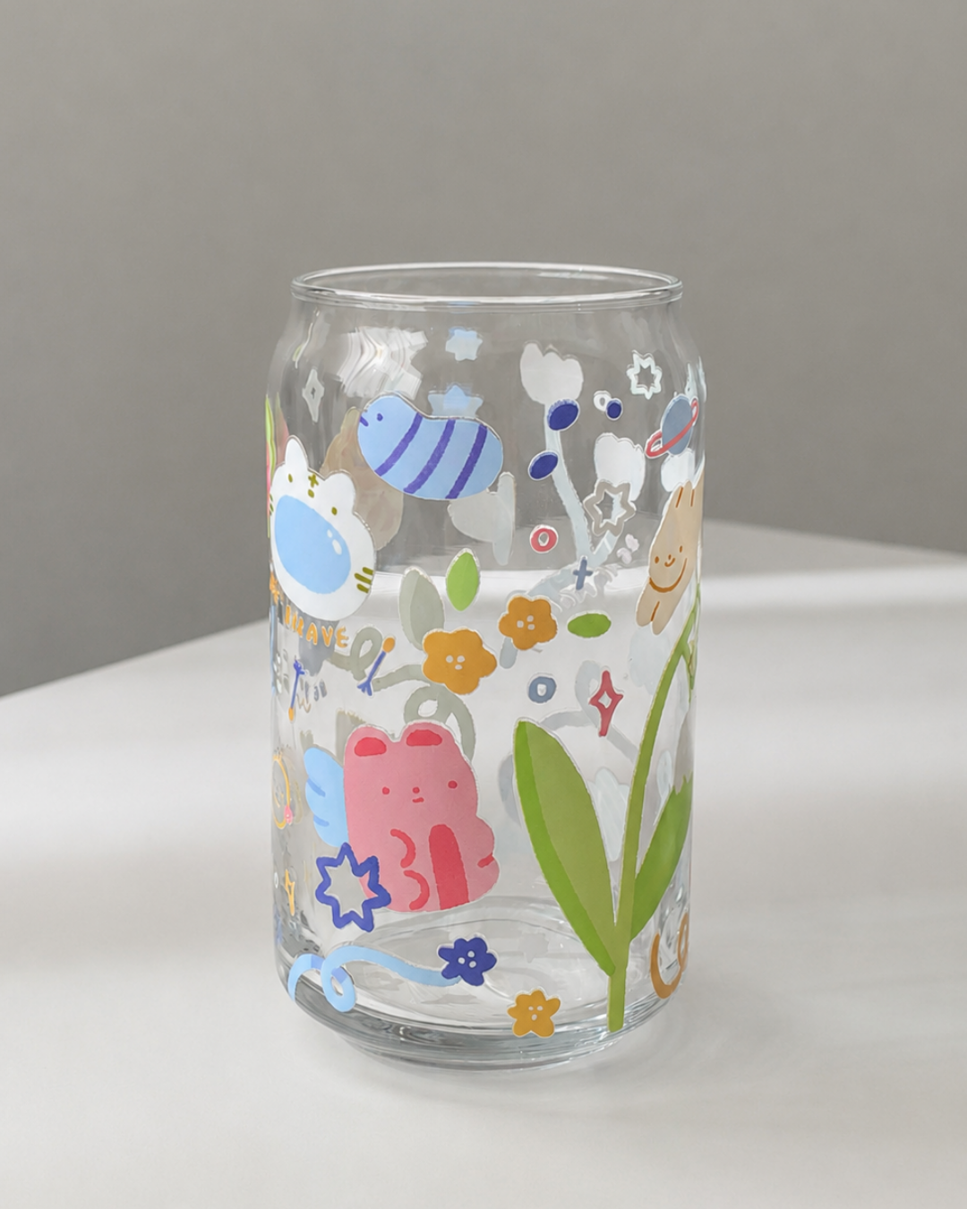 A clear glass can-shaped tumbler decorated with colorful cartoon illustrations, including a pink cat, blue fish, green leaves, and small flowers, set against a neutral gray background.